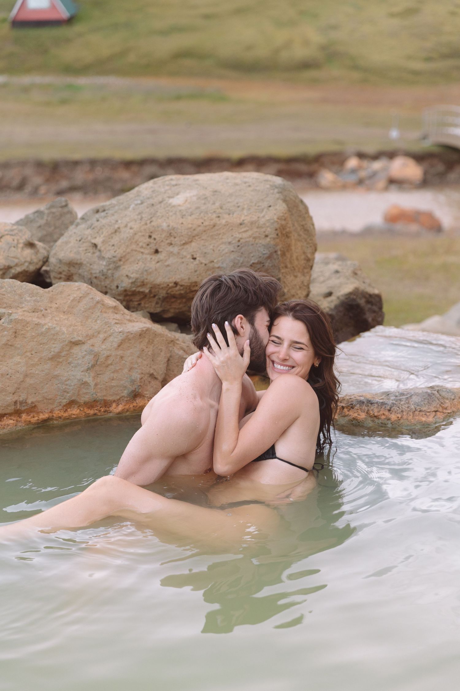 Smiling couple hugging and enjoying a natural geothermal hot spring pool surrounded by large rocks in Iceland.