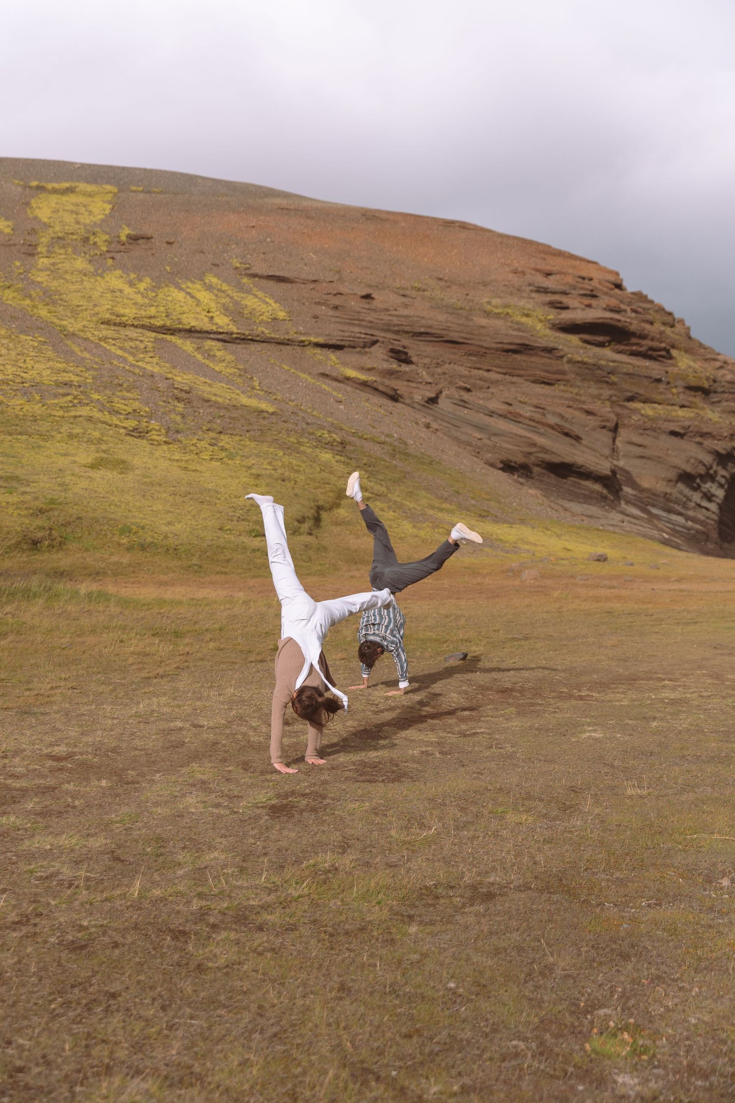 Couple performing simultaneous cartwheels on an open grassy field with colorful rocky cliffs behind.