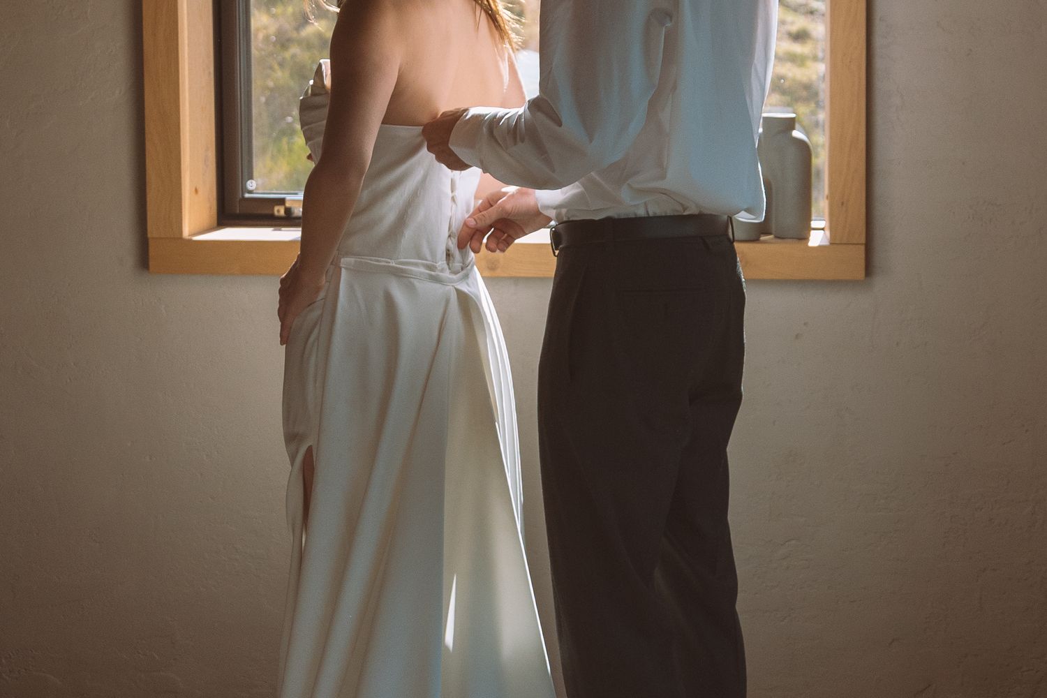 Groom fastening the back of brides elegant white wedding dress near a sunlit window.