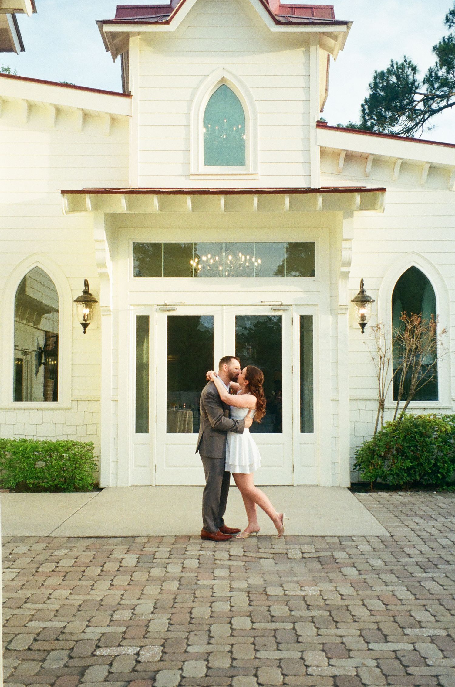 Couple sharing a kiss in front of a charming white chapel with gothic windows.