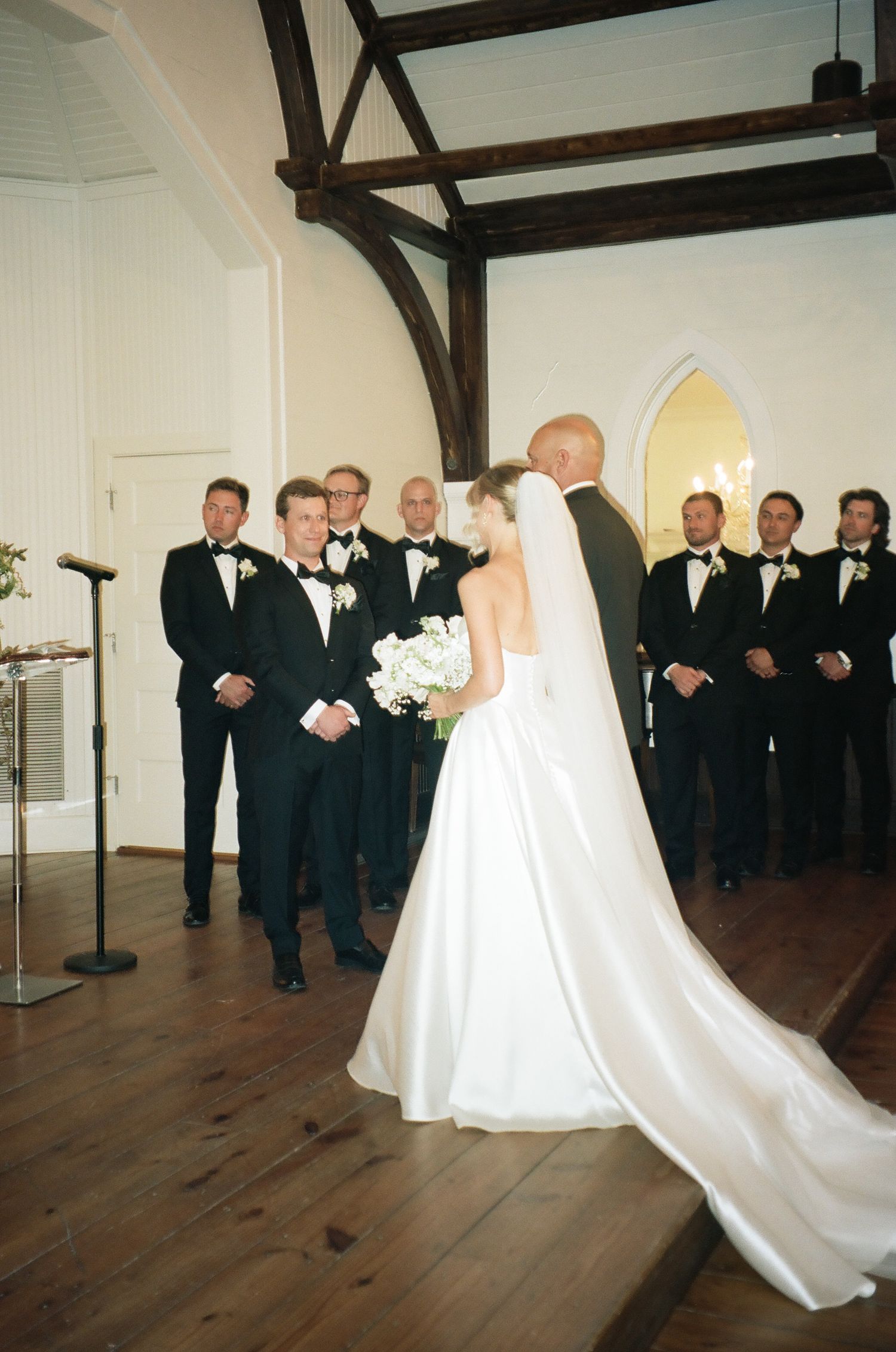 Bride walks down the aisle in a white gown past groomsmen in tuxedos during a church wedding ceremony.