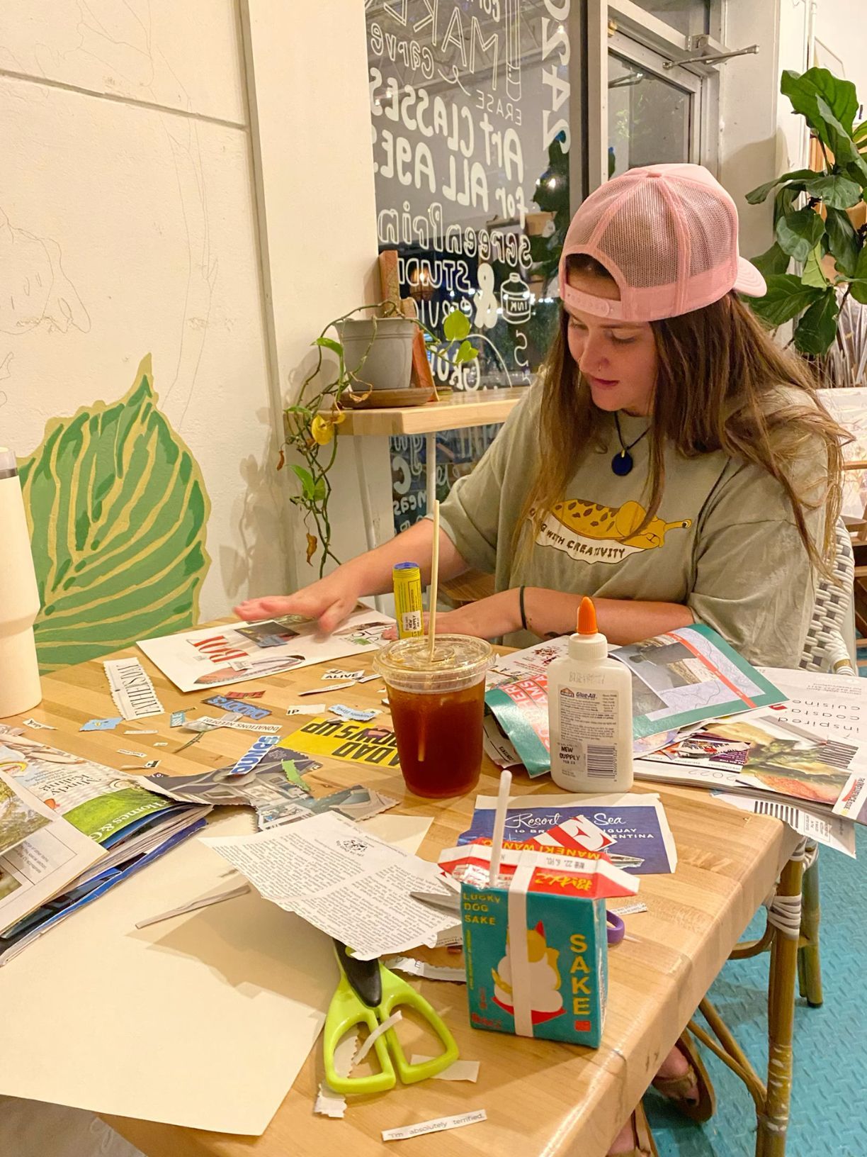 Young woman in a pink cap creating a collage with magazines, scissors, and glue at a craft table.