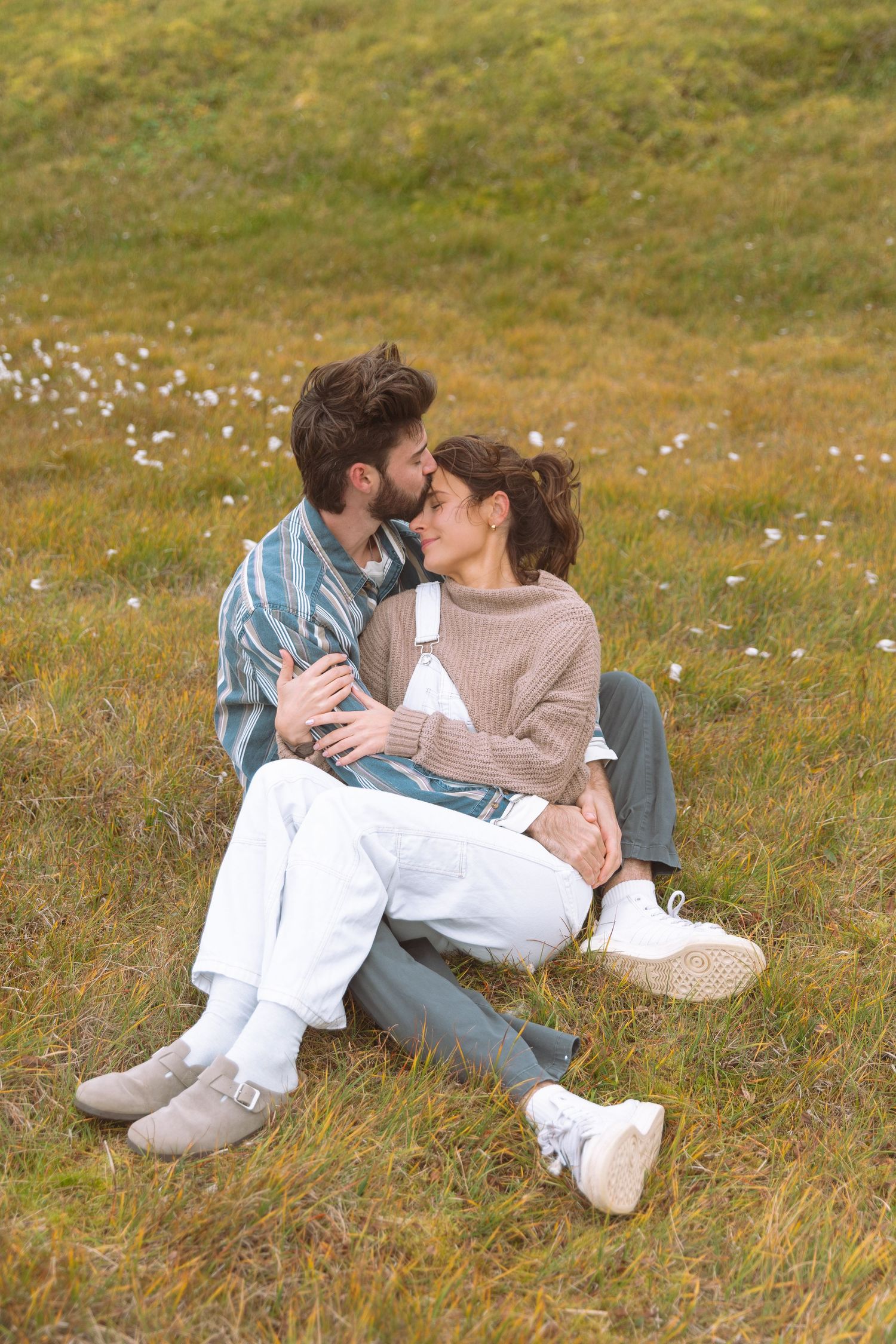 Romantic couple sitting closely together on a grassy hillside, sharing an intimate moment outdoors.