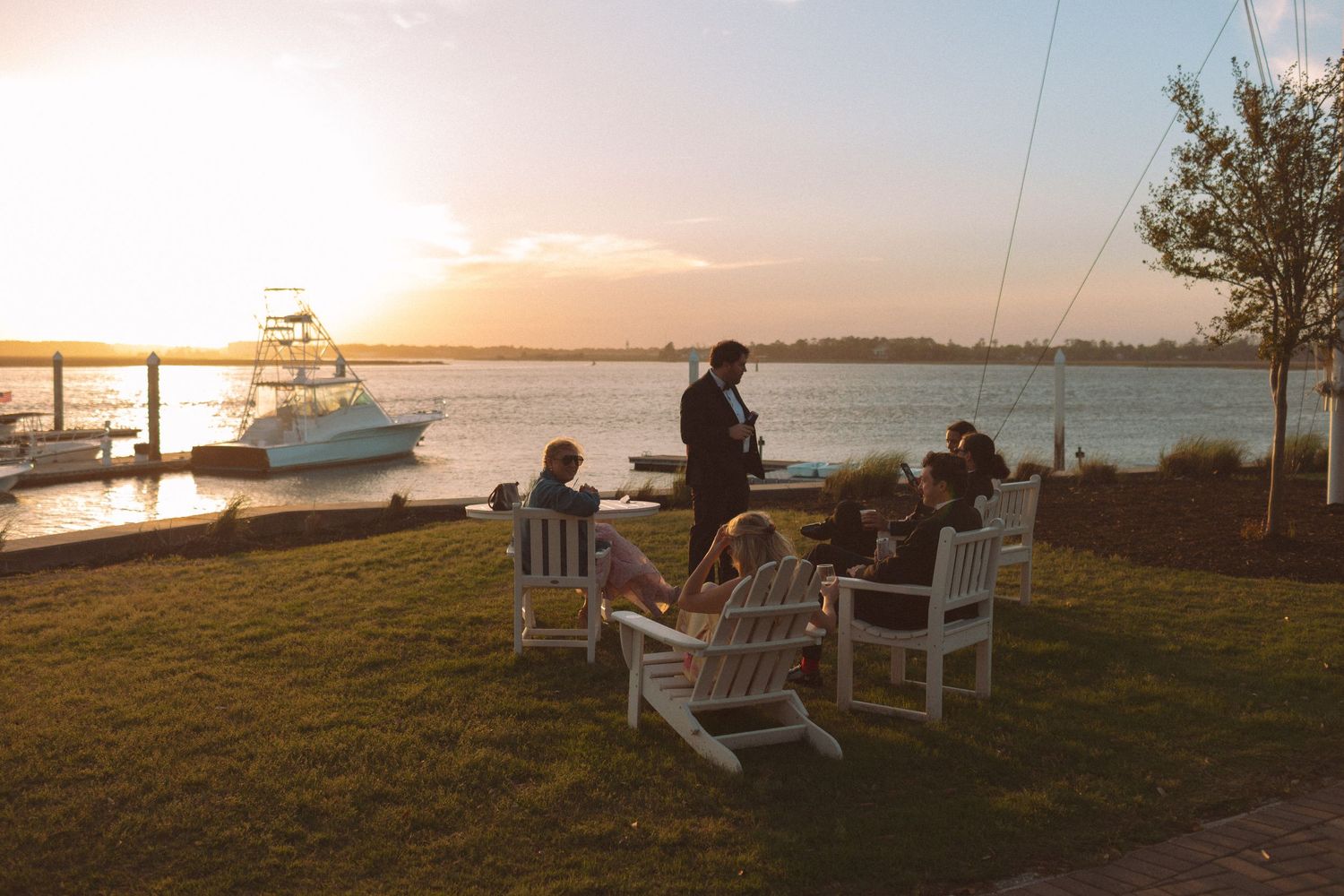 Wedding guests relax in white Adirondack chairs by the waterfront at golden hour sunset with boats docked in the background.