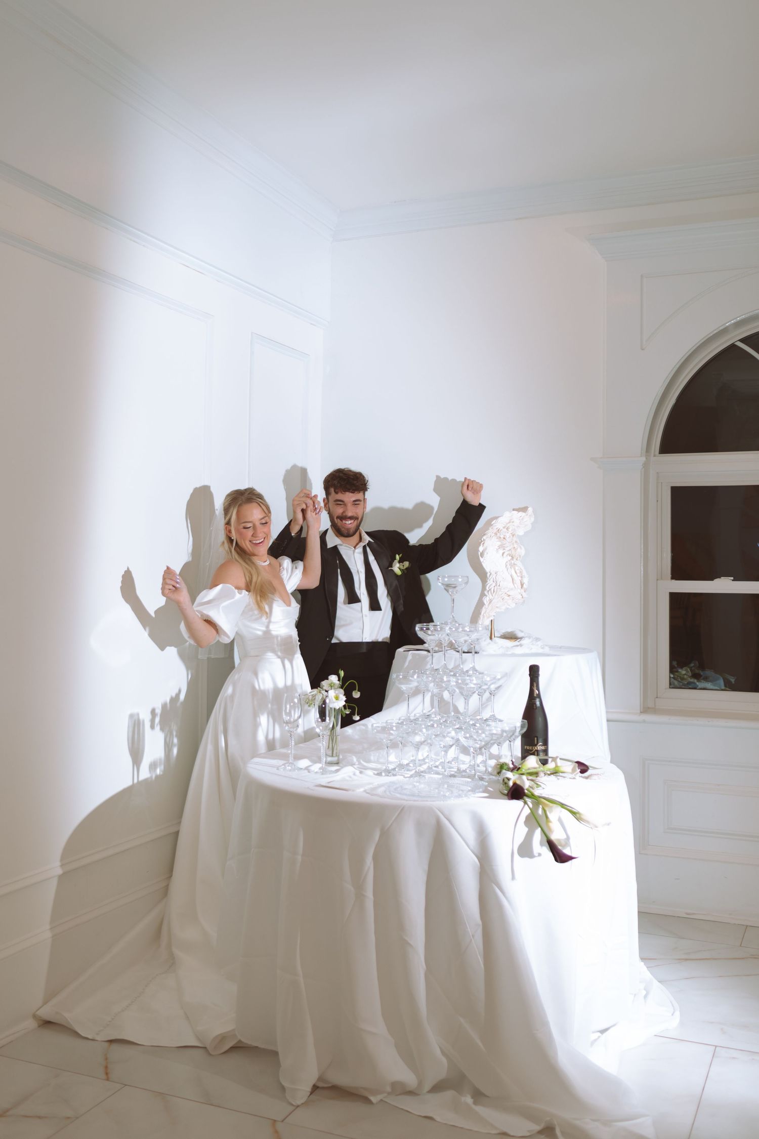 Joyful bride and groom celebrating at champagne tower table in elegant white wedding venue.