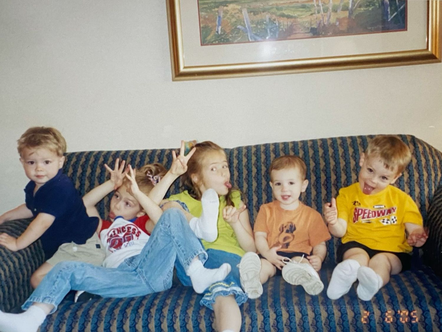 Five young children sitting together on a blue sofa, making silly faces and gestures at the camera.