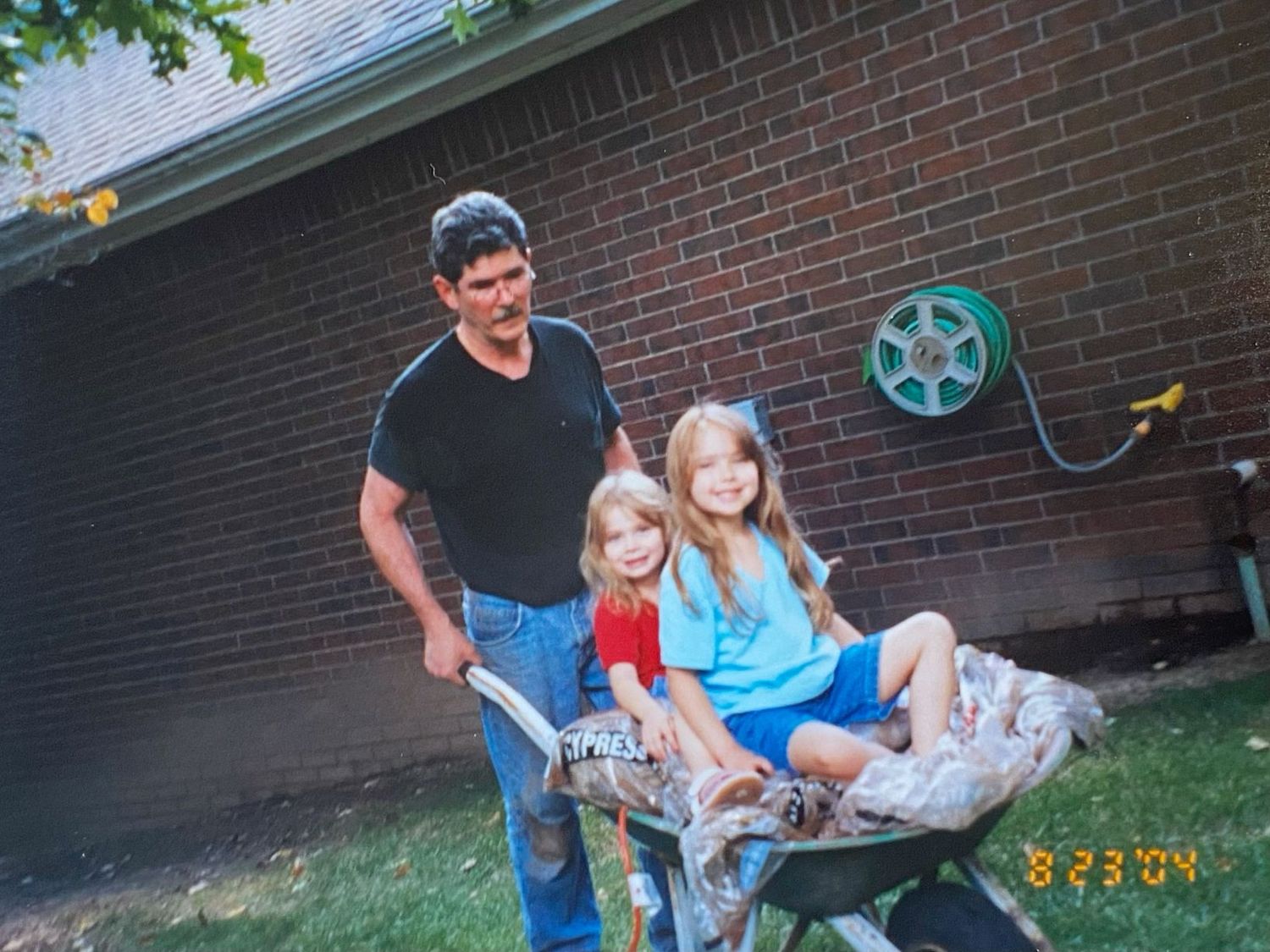 Father pushing two young girls in a wheelbarrow in a backyard beside a brick wall.