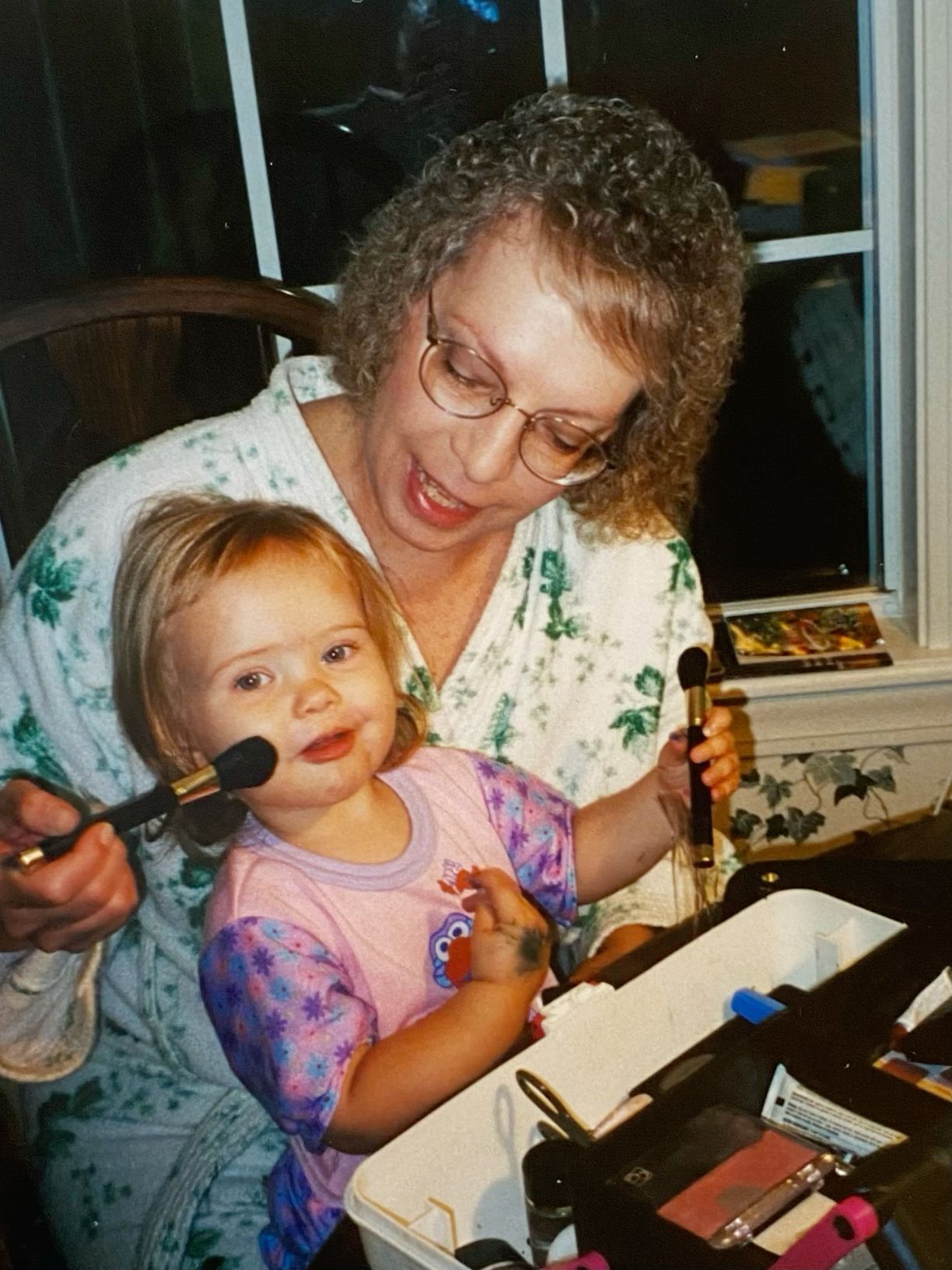 A curly-haired grandmother applies makeup with a brush on a smiling toddler girl.
