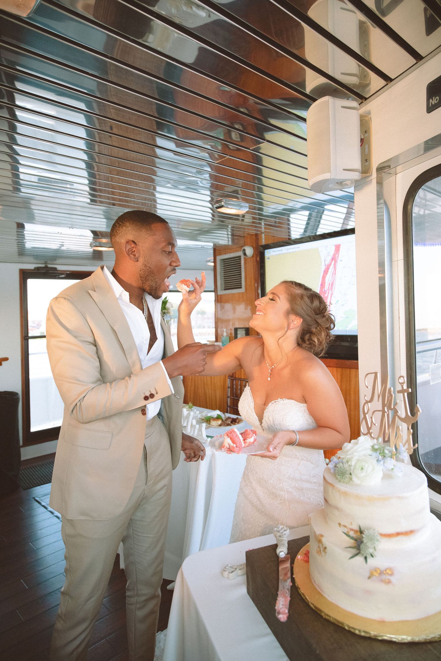Groom in beige suit feeds wedding cake to bride in white strapless gown during reception on a boat.