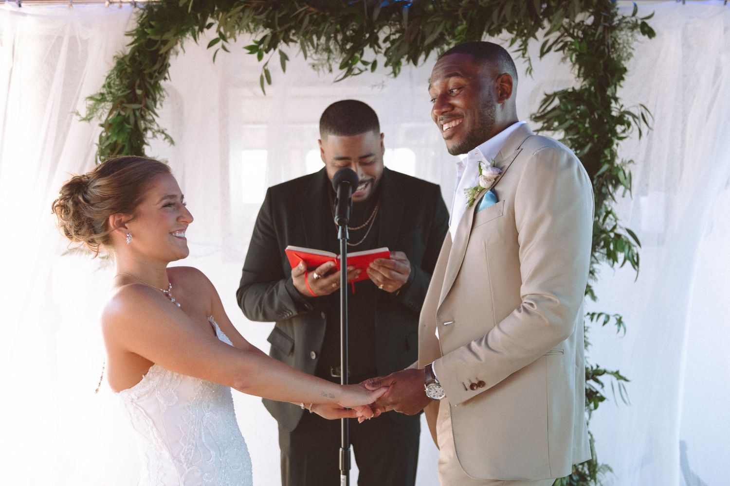 Bride and groom exchange vows and laugh joyfully during wedding ceremony under greenery arch.