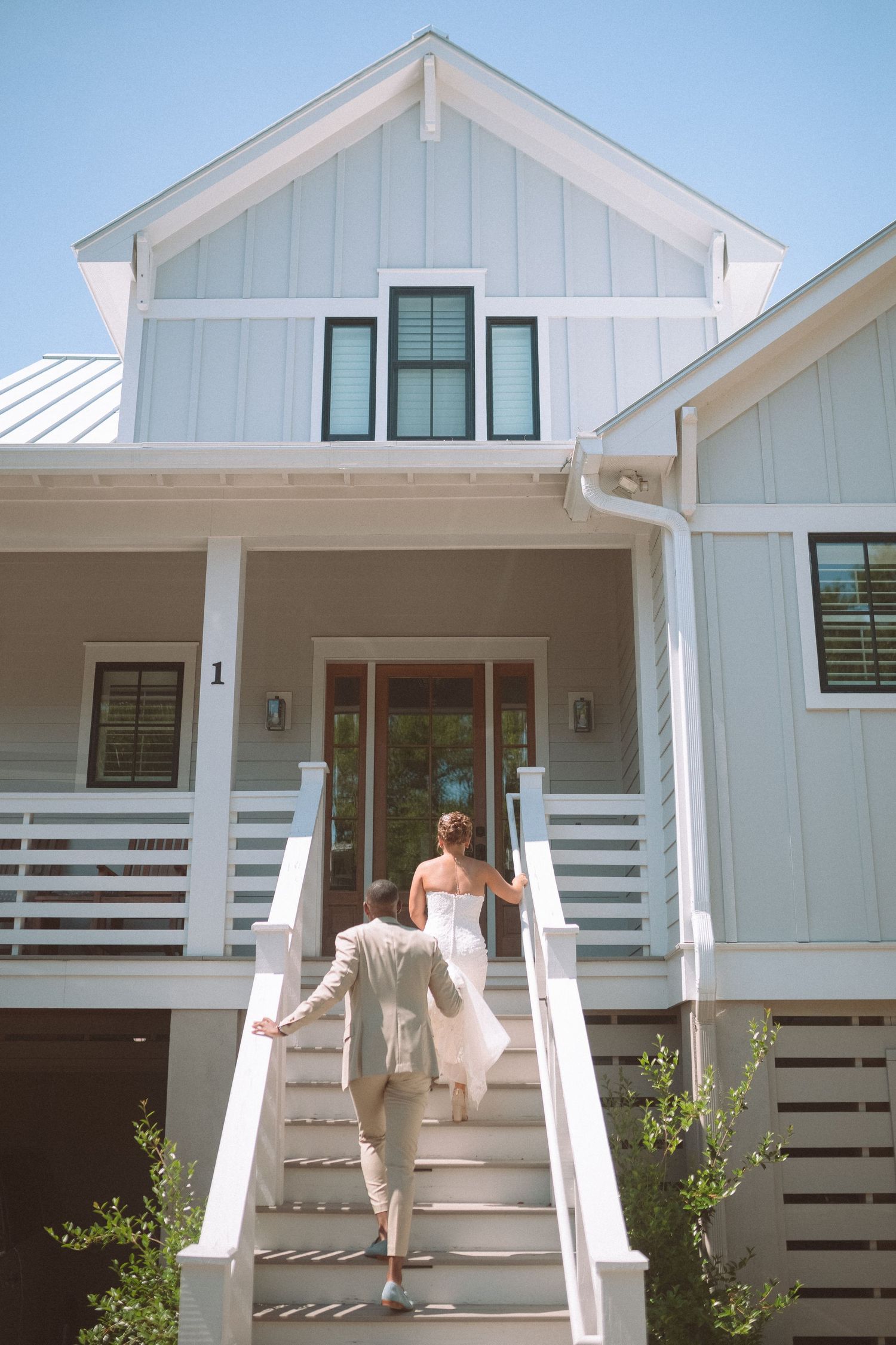 Bride and groom walking up white stairs toward charming coastal farmhouse on wedding day.