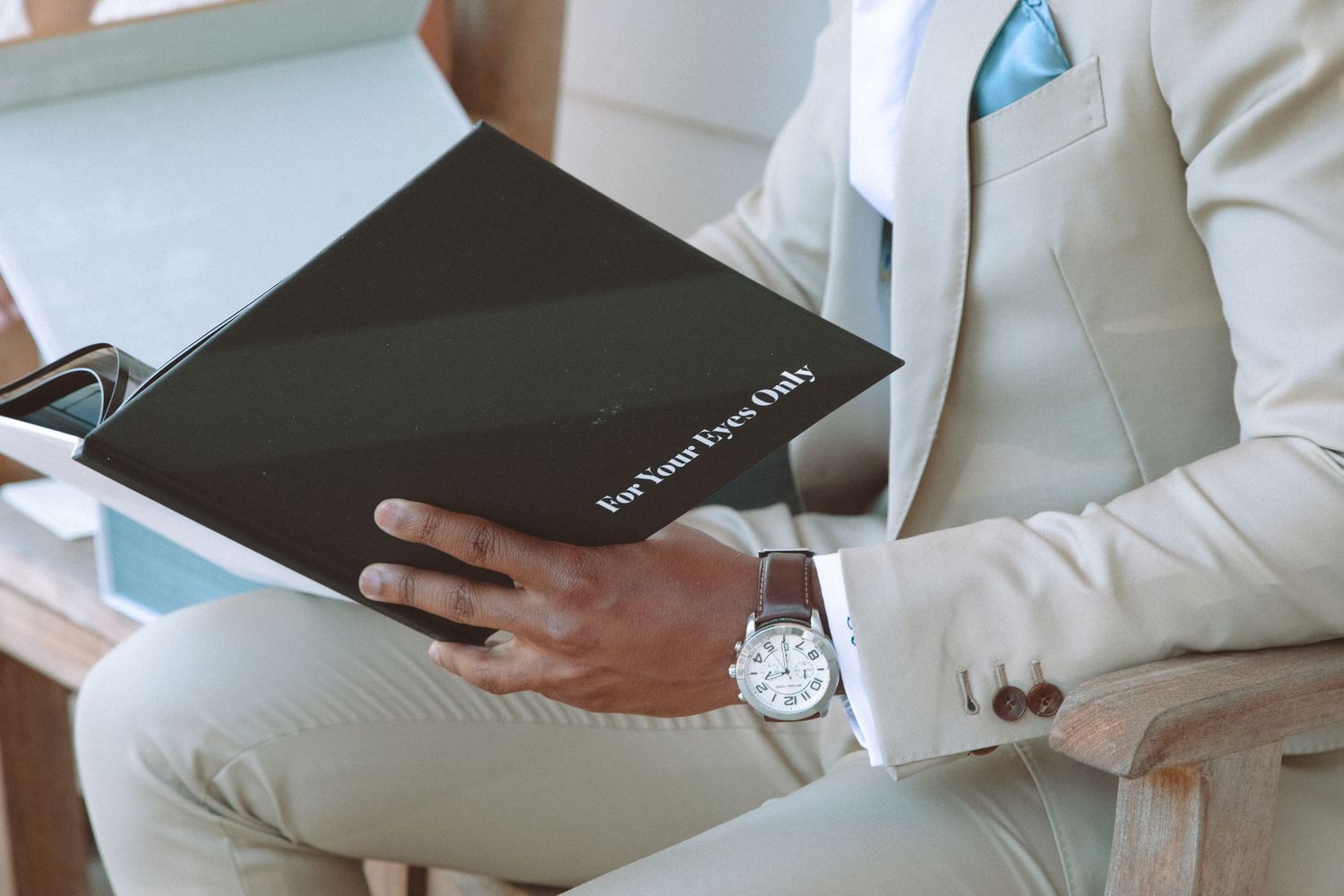 Close-up of groom in beige suit holding black wedding album inscribed with For Your Eyes Only.