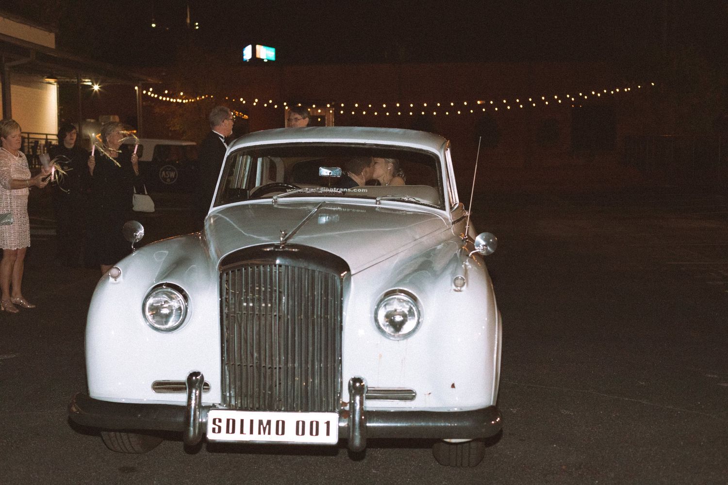 Bride and groom depart wedding reception in classic vintage white Bentley limousine at night.