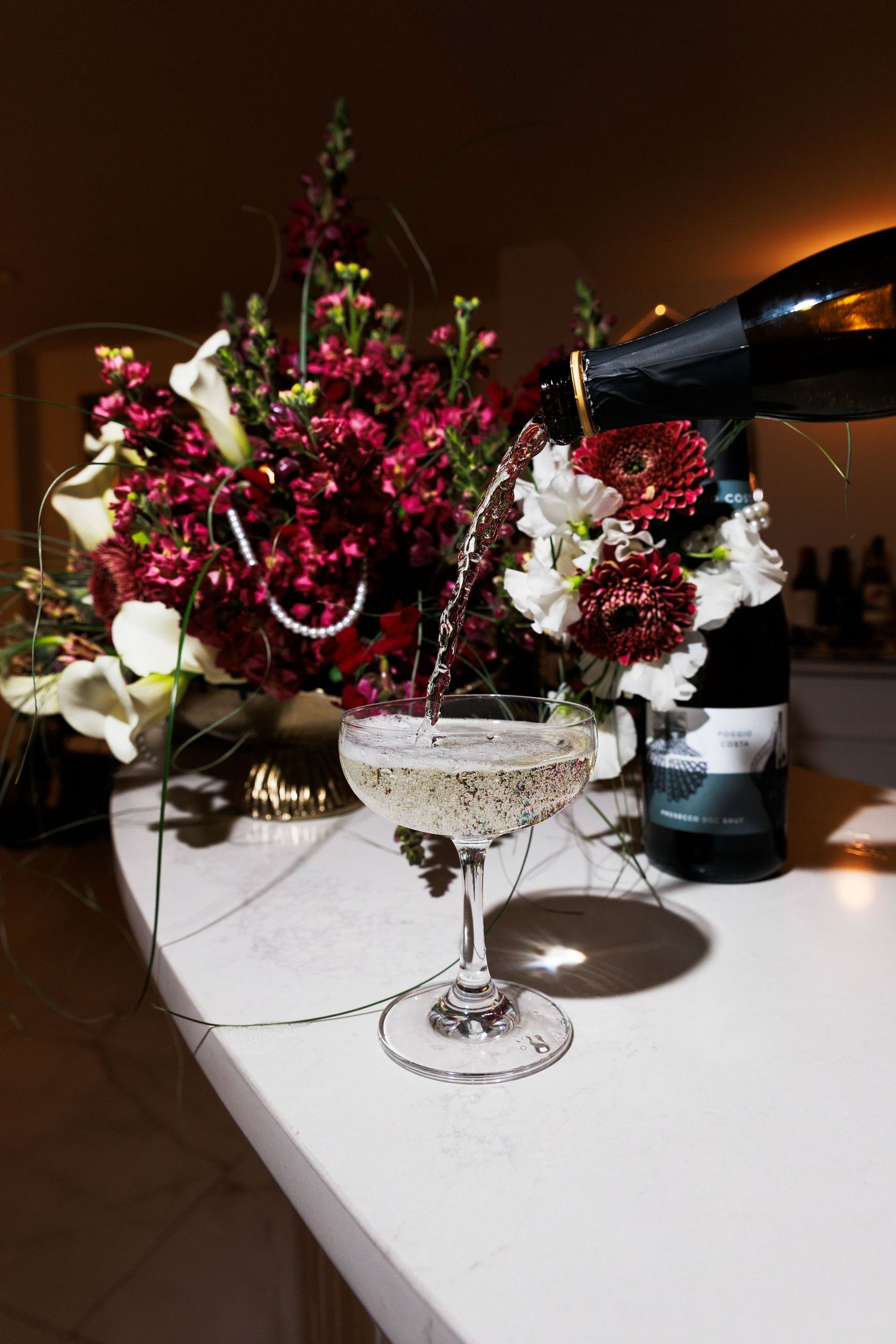 Champagne being poured into a coupe glass beside a bottle and red floral arrangement on a white marble surface.