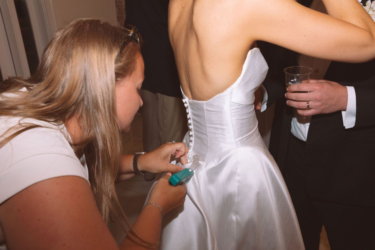Bridesmaid fastening buttons on back of white satin wedding dress during bridal preparation.