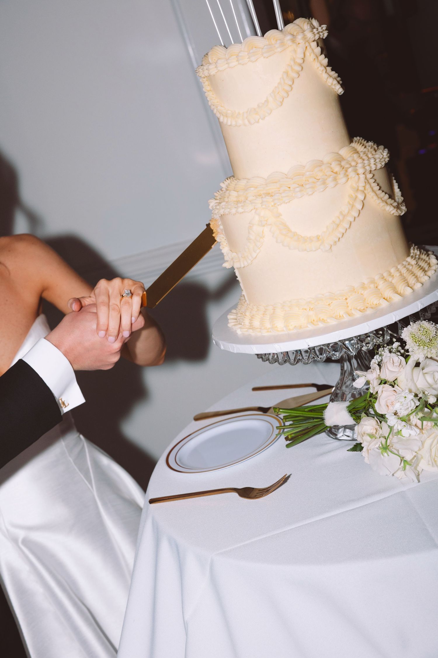 Bride and groom cutting elegant three-tier white wedding cake with pearl and ruffle decorations.