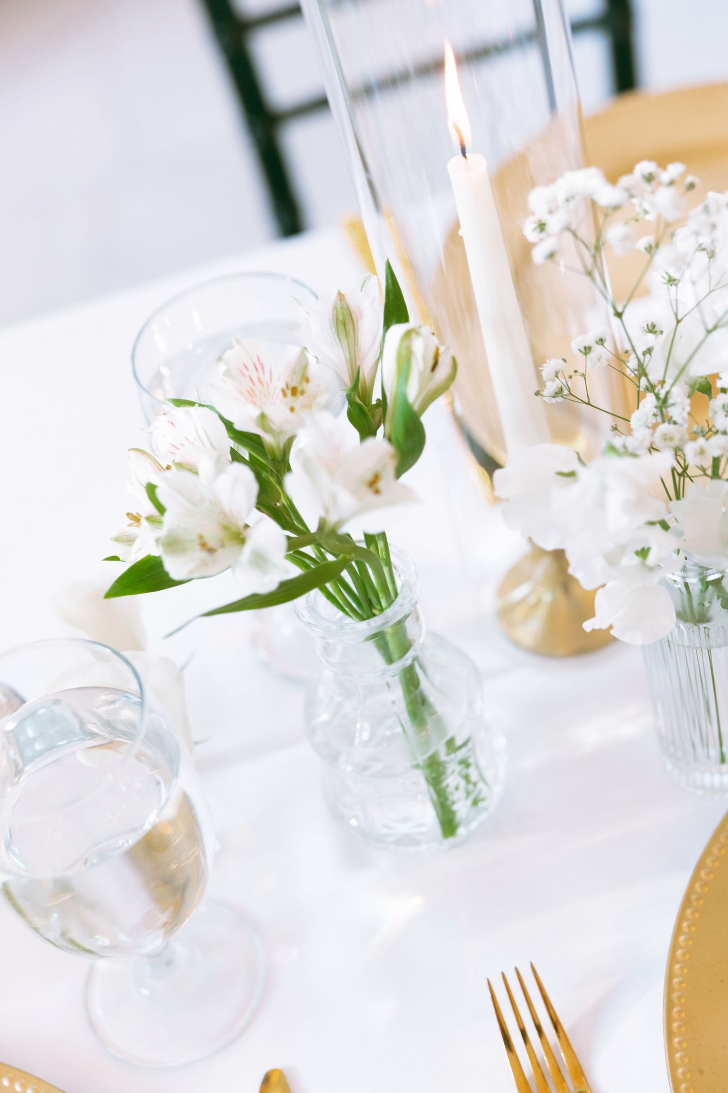 Elegant wedding table setting with white flowers in glass vases, gold cutlery, and lit candles on white linen.