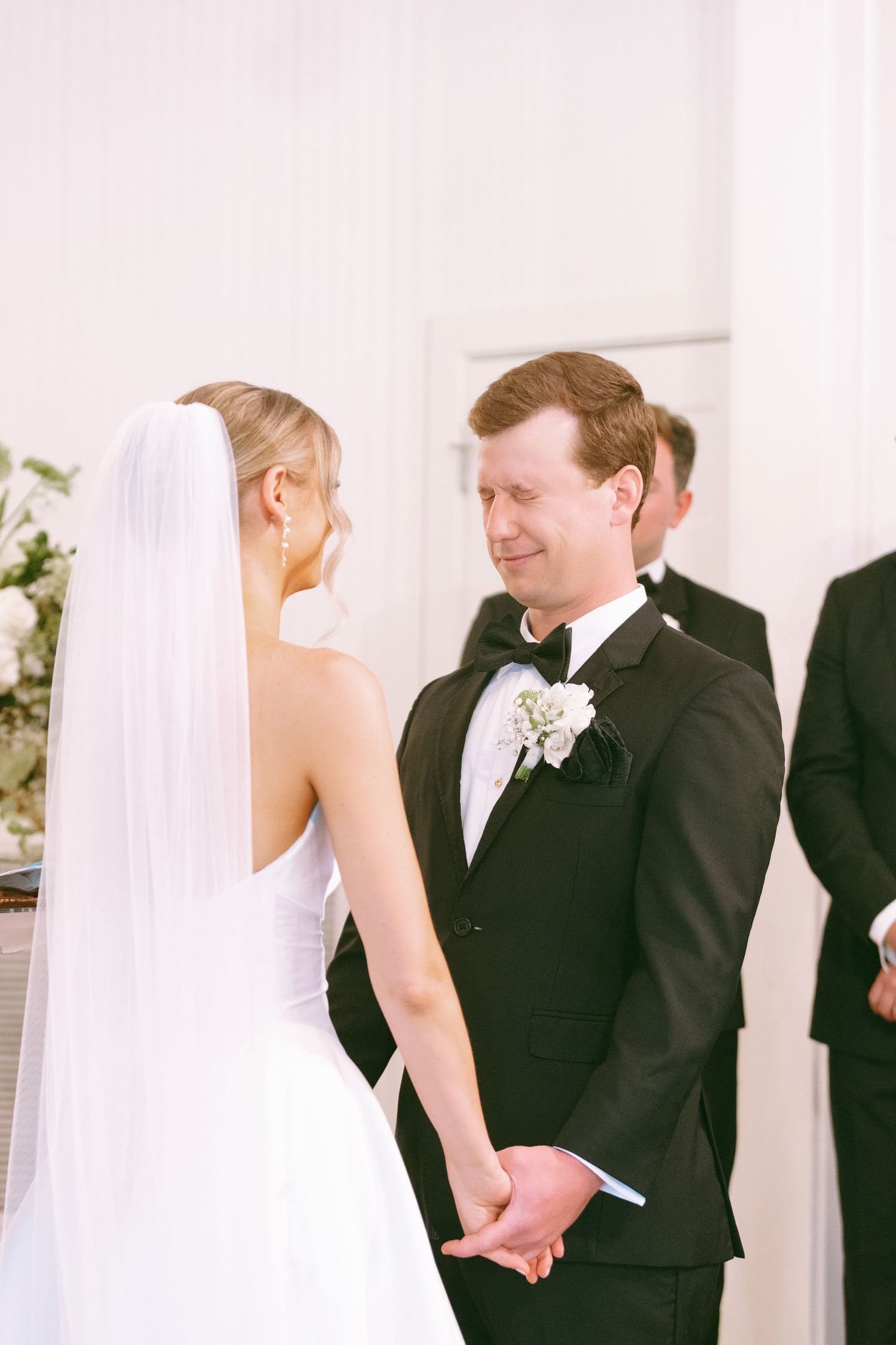Bride and groom hold hands at altar during wedding ceremony, groom smiling with white floral boutonniere.