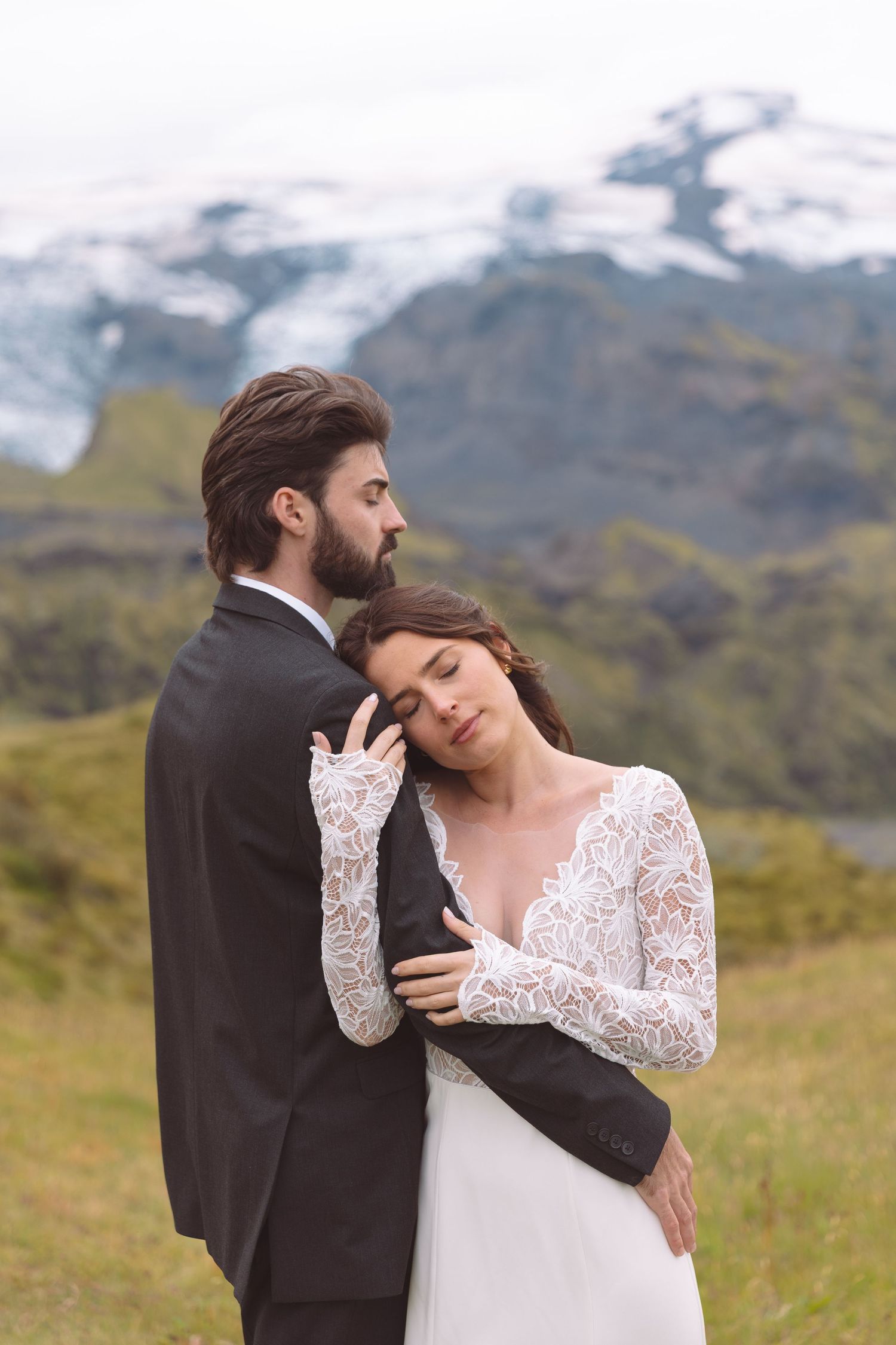 Bride in lace wedding dress rests head on grooms chest in scenic mountain landscape.