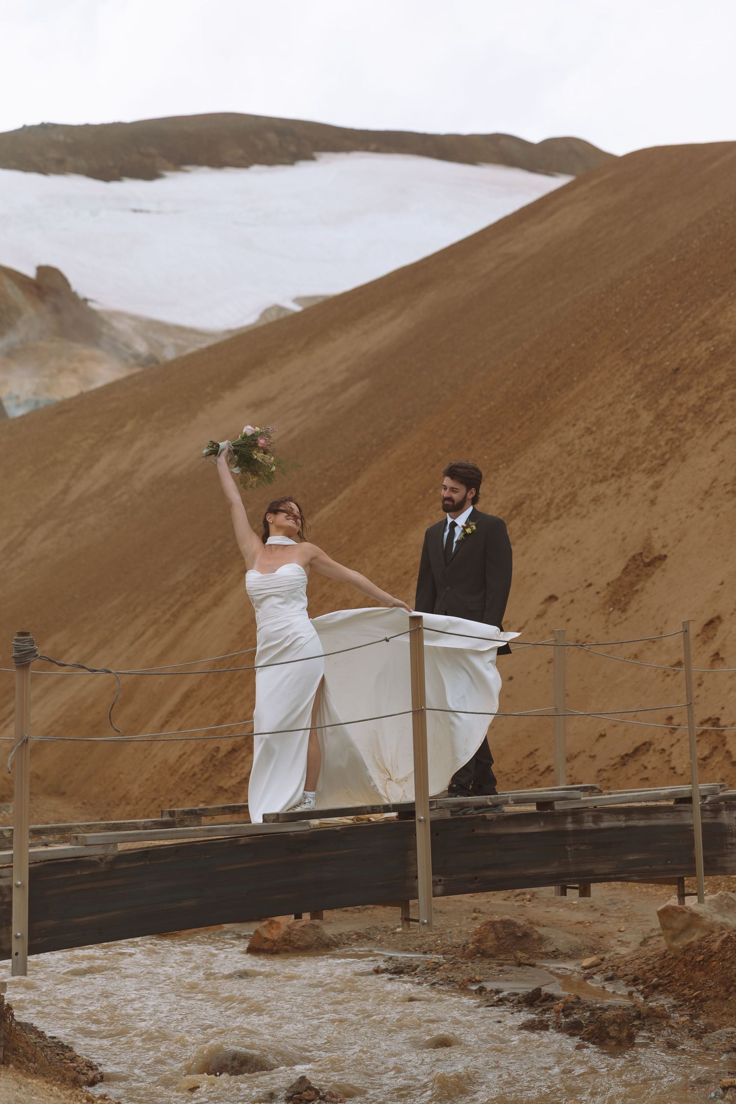 Bride raises bouquet joyfully as groom smiles on a wooden bridge against dramatic Iceland mountains.