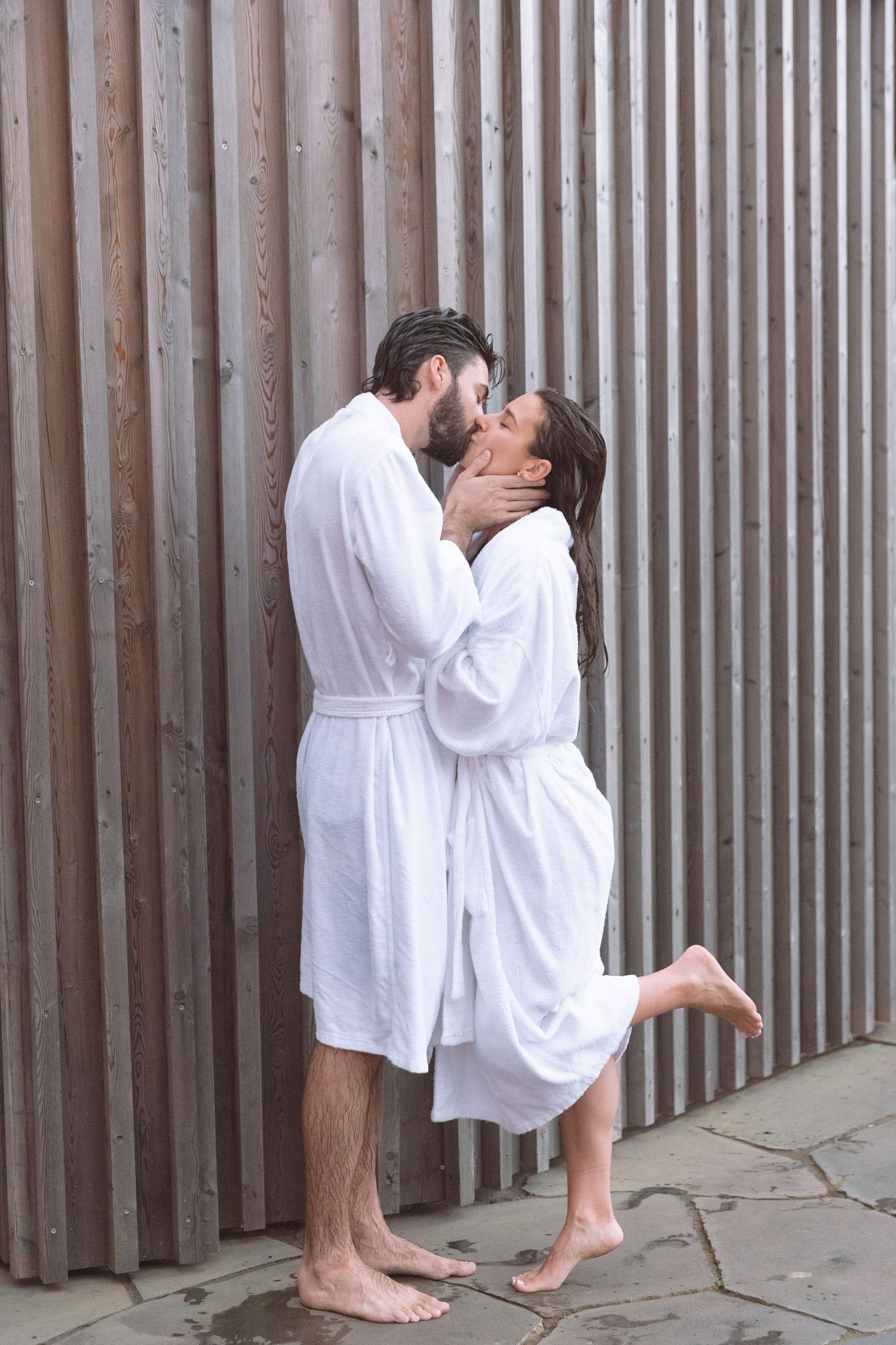 Couple in white bathrobes sharing passionate kiss against wooden wall at outdoor spa.