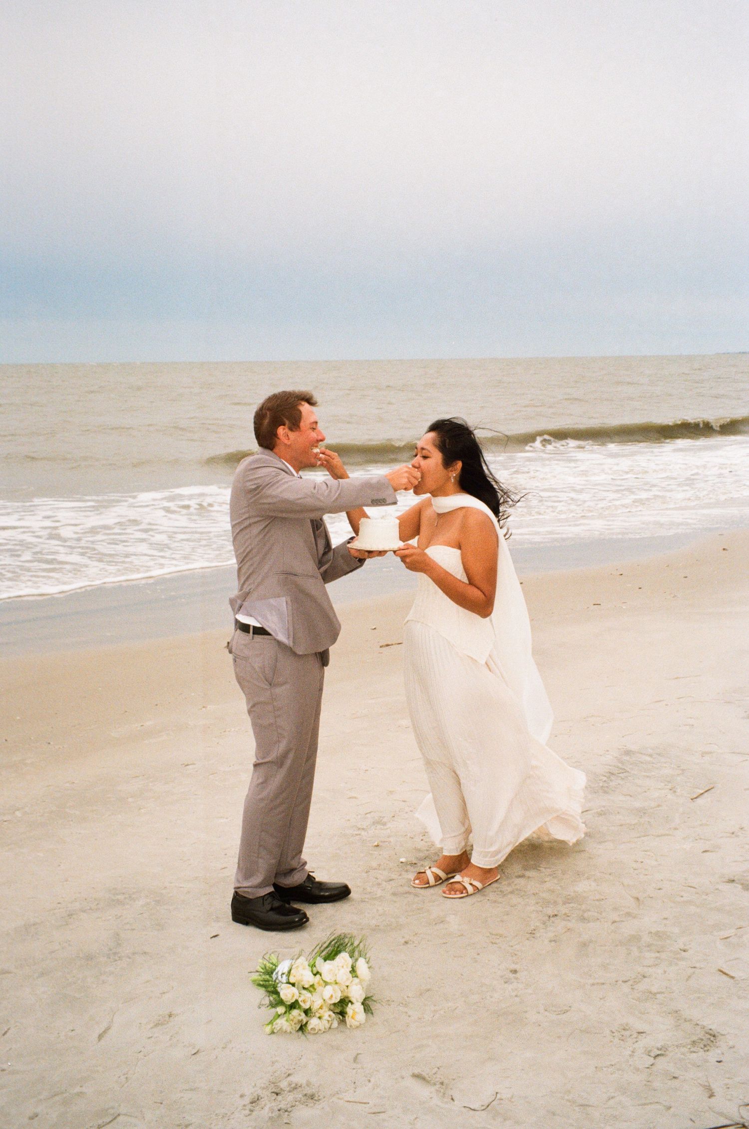 Bride and groom sharing wedding cake on a sandy beach with ocean waves in the background.