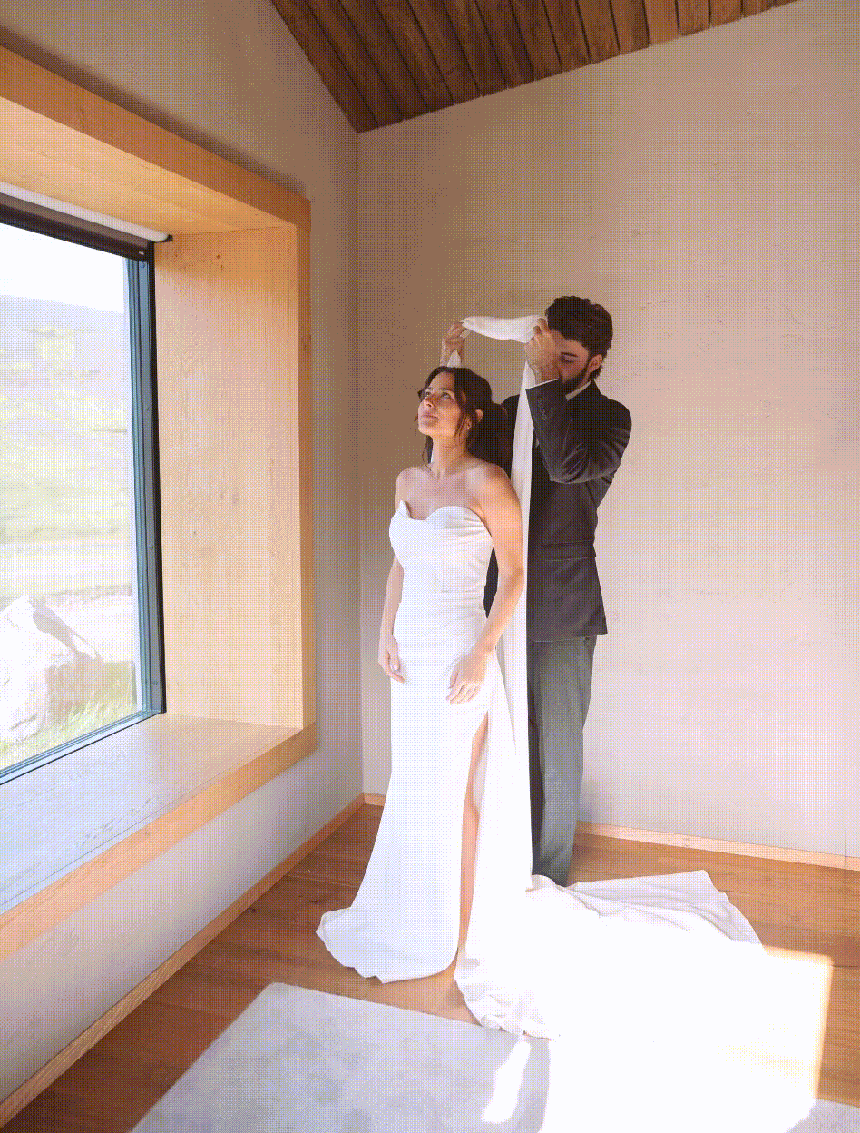 Groom placing veil on bride in white wedding dress standing by window in softly lit room.