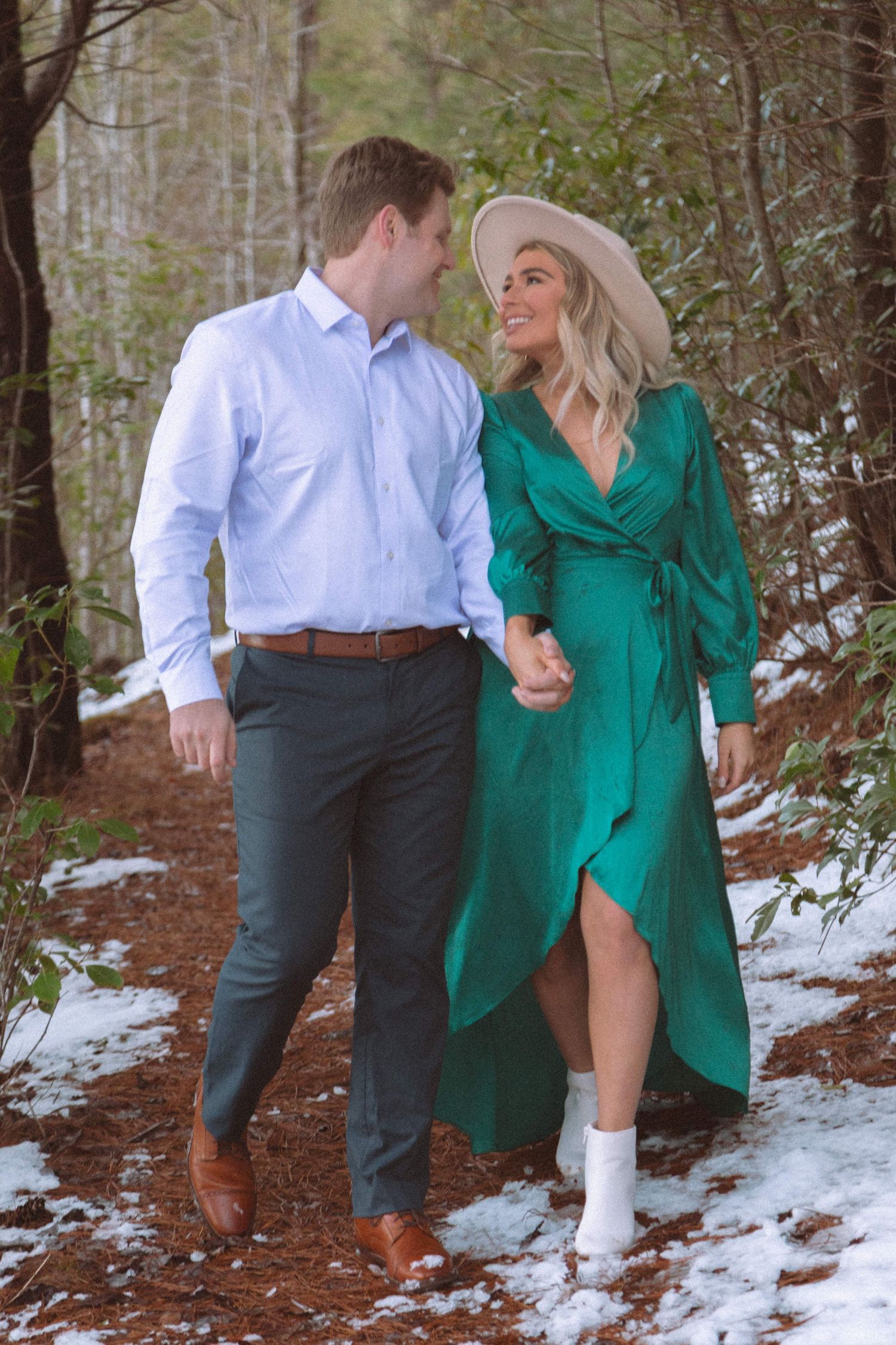 Couple walking hand in hand through snowy forest, woman in elegant green wrap dress.