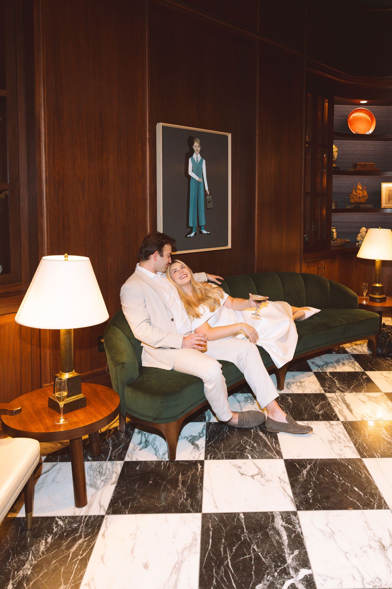 Couple relaxing on a green velvet sofa in an elegant wood-paneled room with checkered marble floors and warm lighting.