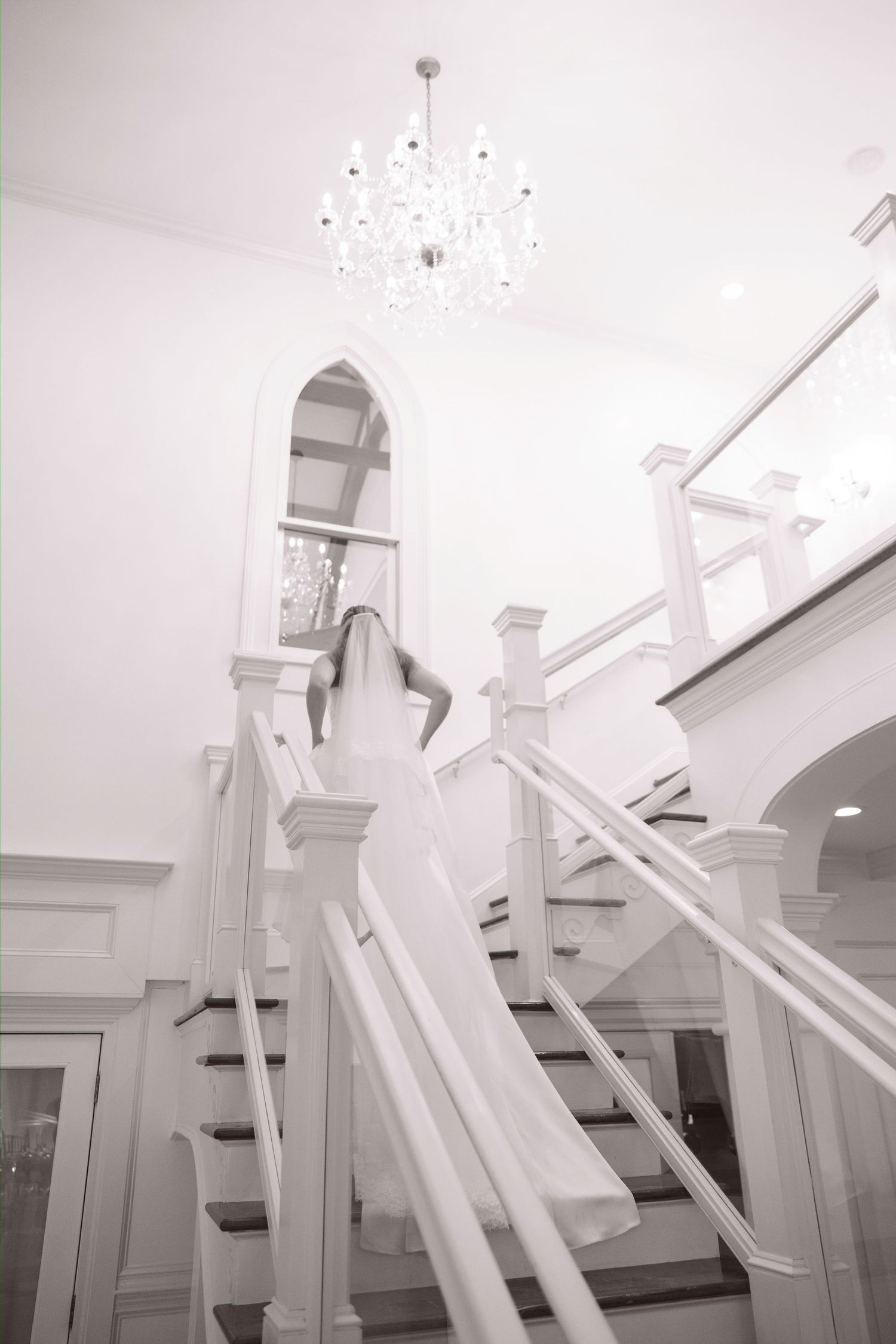 Bride in white gown ascending elegant white staircase beneath a crystal chandelier.