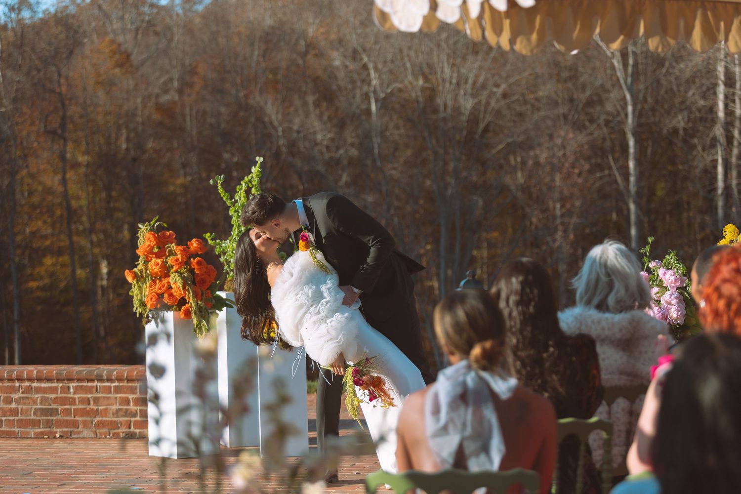 Groom dipping bride at outdoor fall wedding ceremony surrounded by vibrant orange floral arrangements.