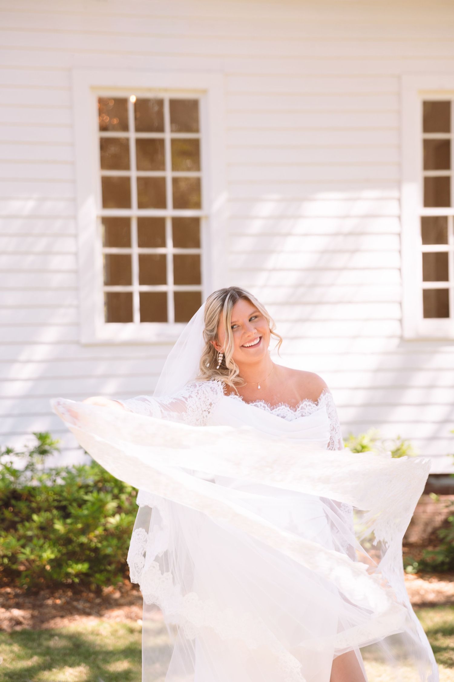 Smiling bride in white lace off-shoulder gown twirling veil outdoors near white building on sunny day.