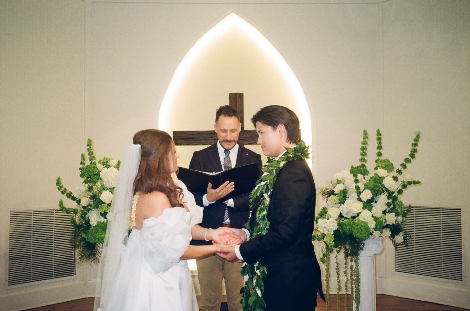 Bride and groom exchange vows at a church altar with a officiant, floral arrangements, and a cross backdrop.