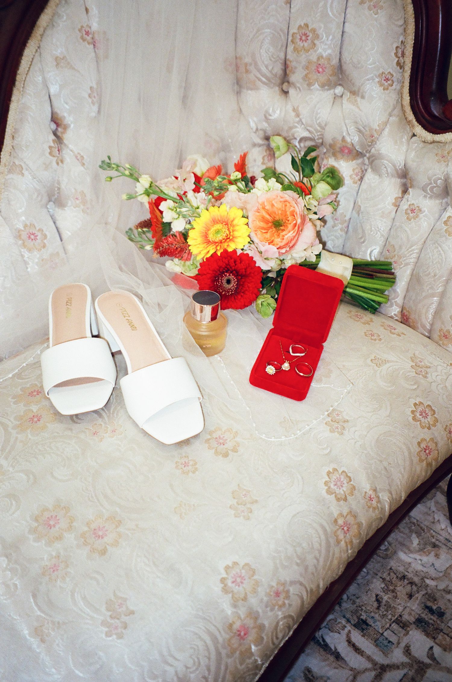Bridal flat shoes, colorful bouquet, perfume bottle, and jewelry in red box arranged on vintage floral chair.