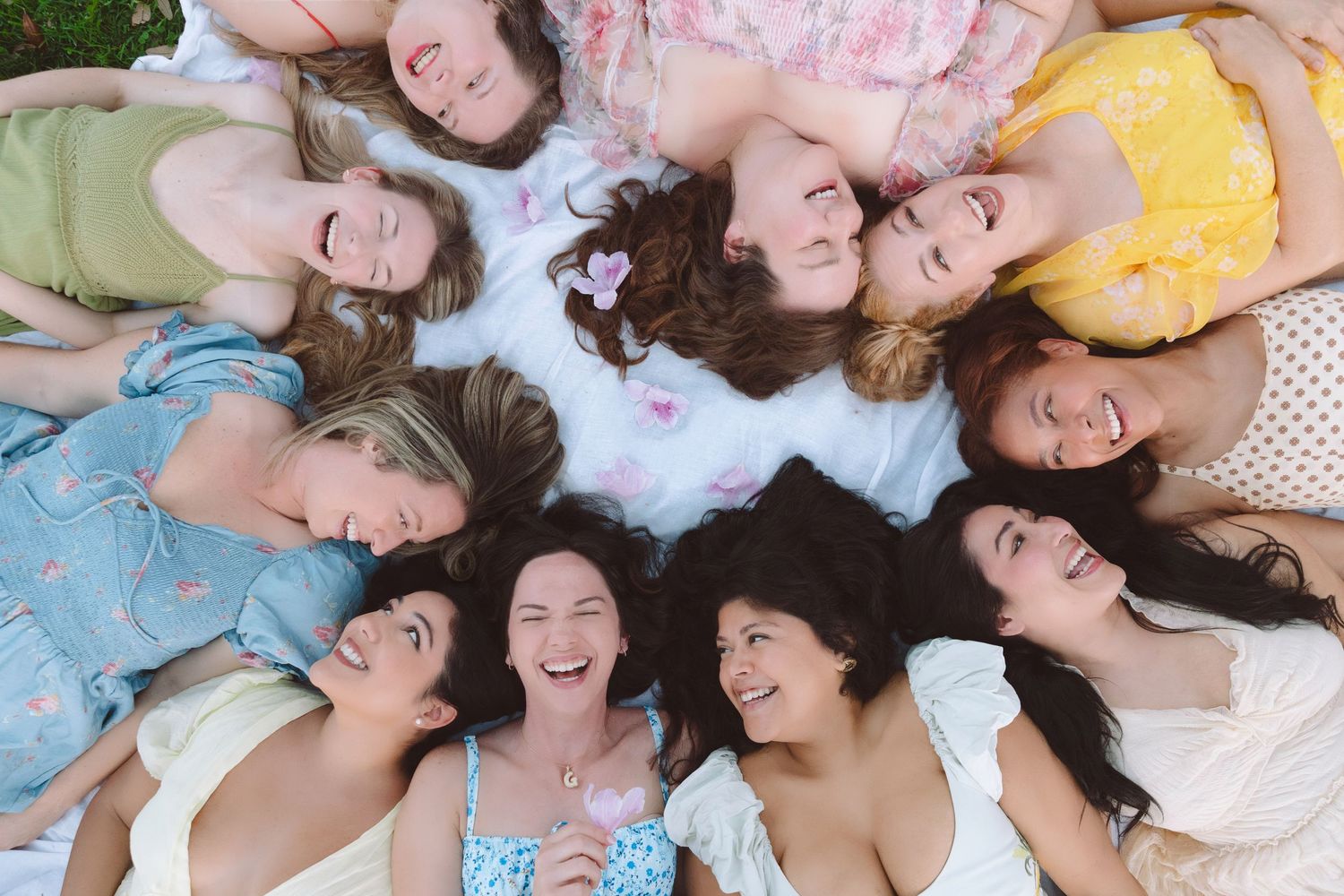 Group of smiling women lying in a circle on the ground, heads together, enjoying a fun outdoor gathering.