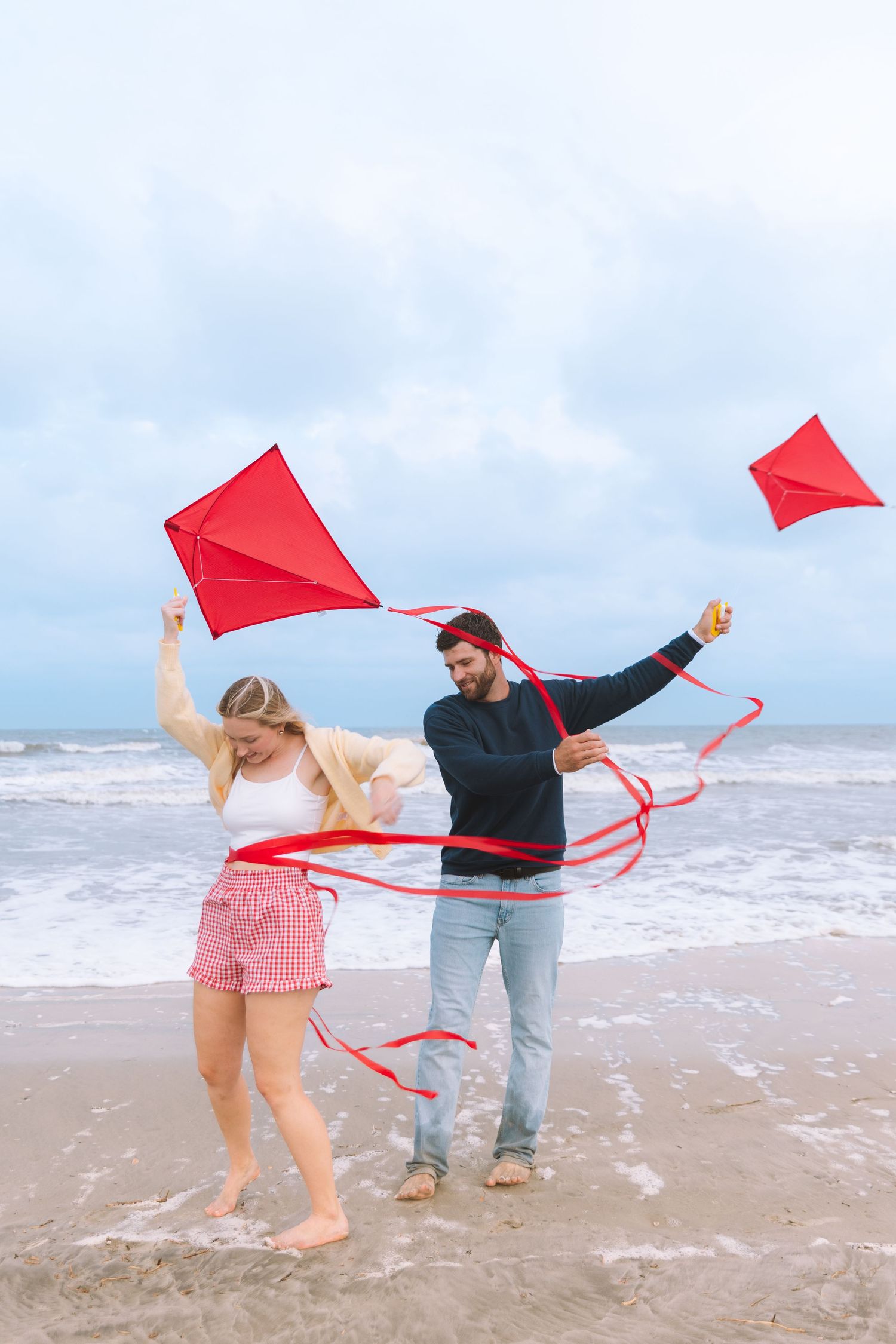 Couple laughing and playing with two red kites tangled in the wind on a beach.