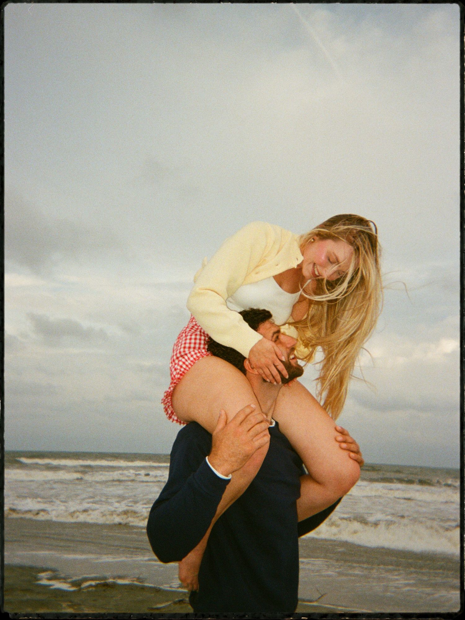 Man carrying woman on his back at beach with waves and cloudy sky behind them.