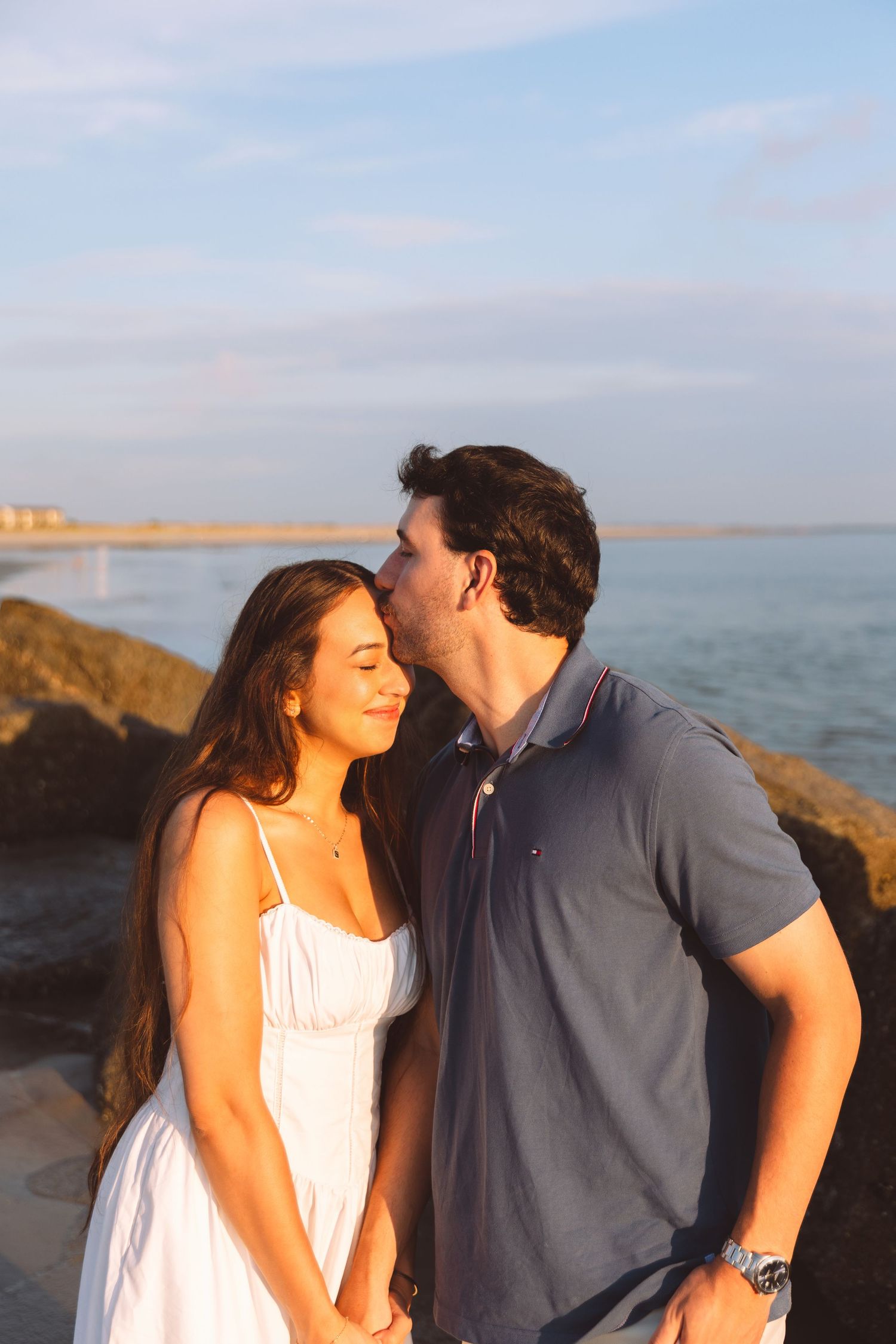 Man kissing woman on the forehead at golden hour on a rocky beach, woman smiling in white sundress.