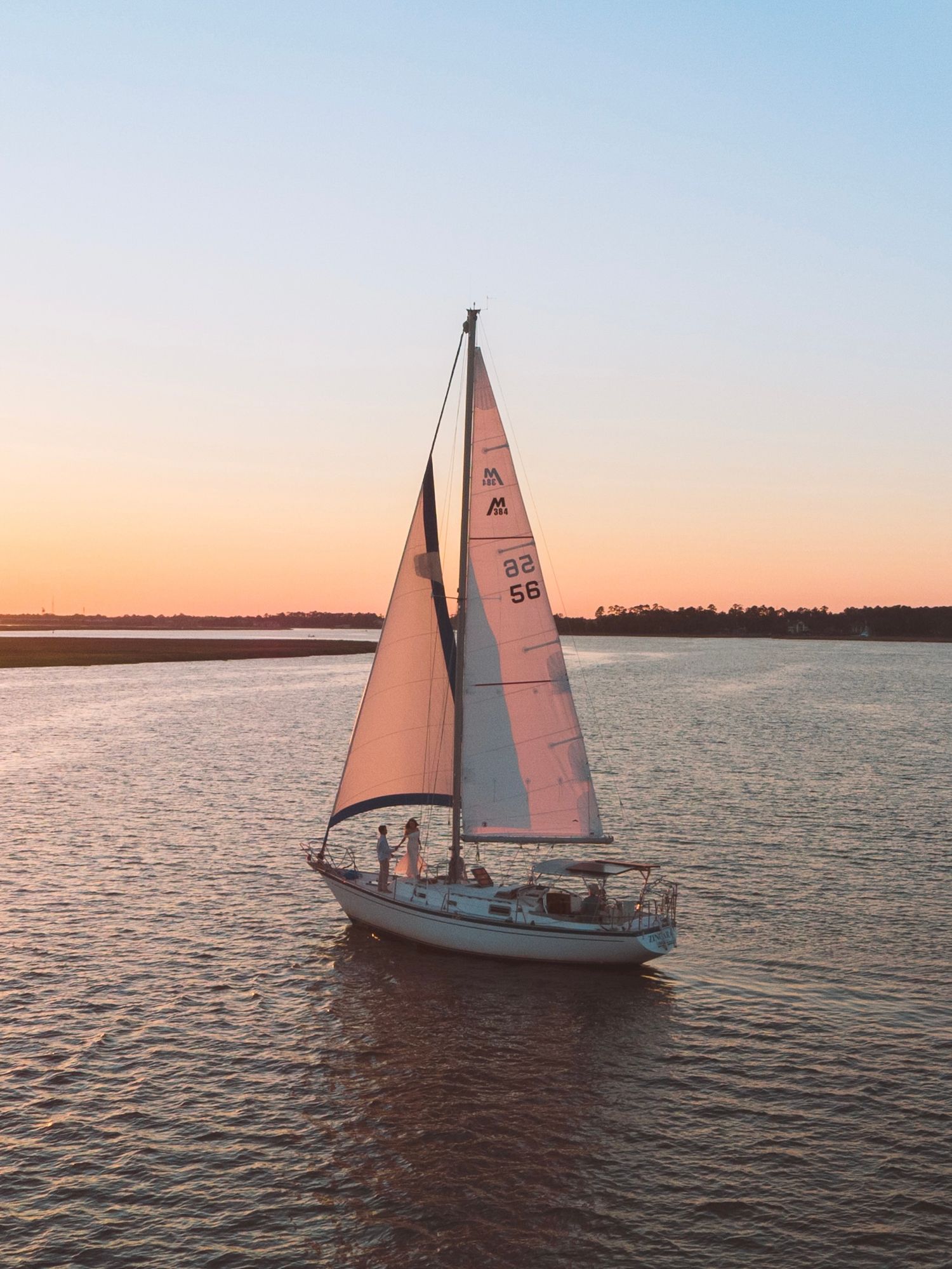 Sailboat with white sails gliding on calm water during a golden sunset.