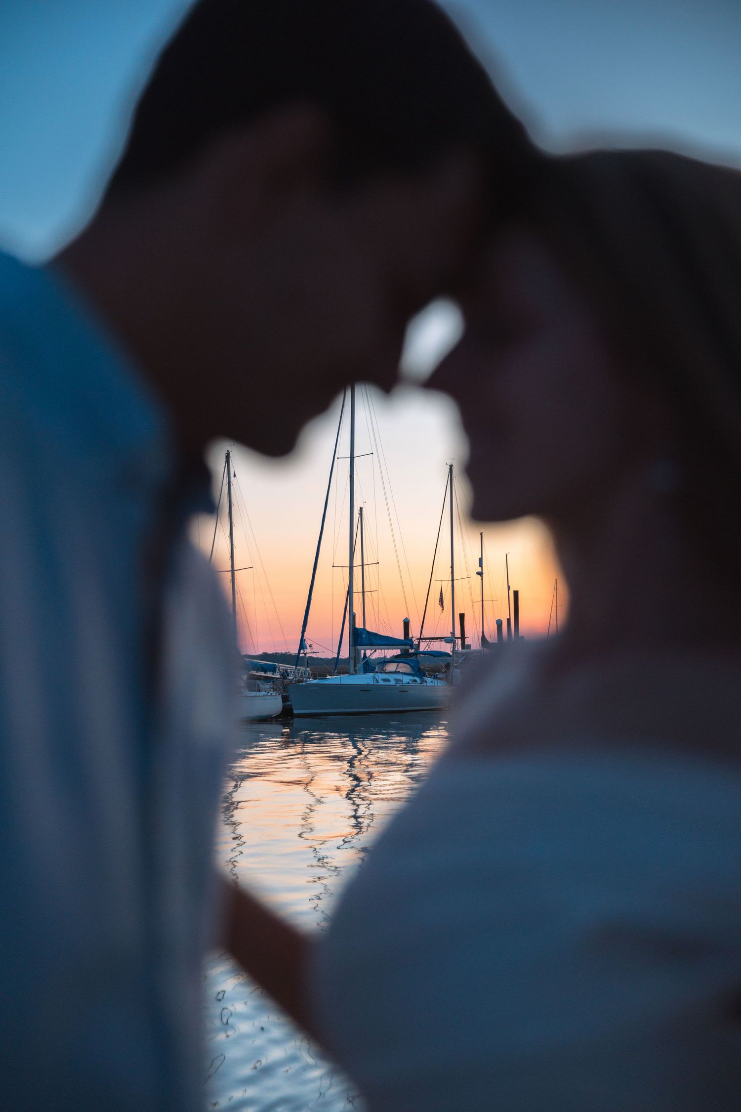 Romantic couple silhouetted against a stunning marina sunset with sailboats reflected in water.
