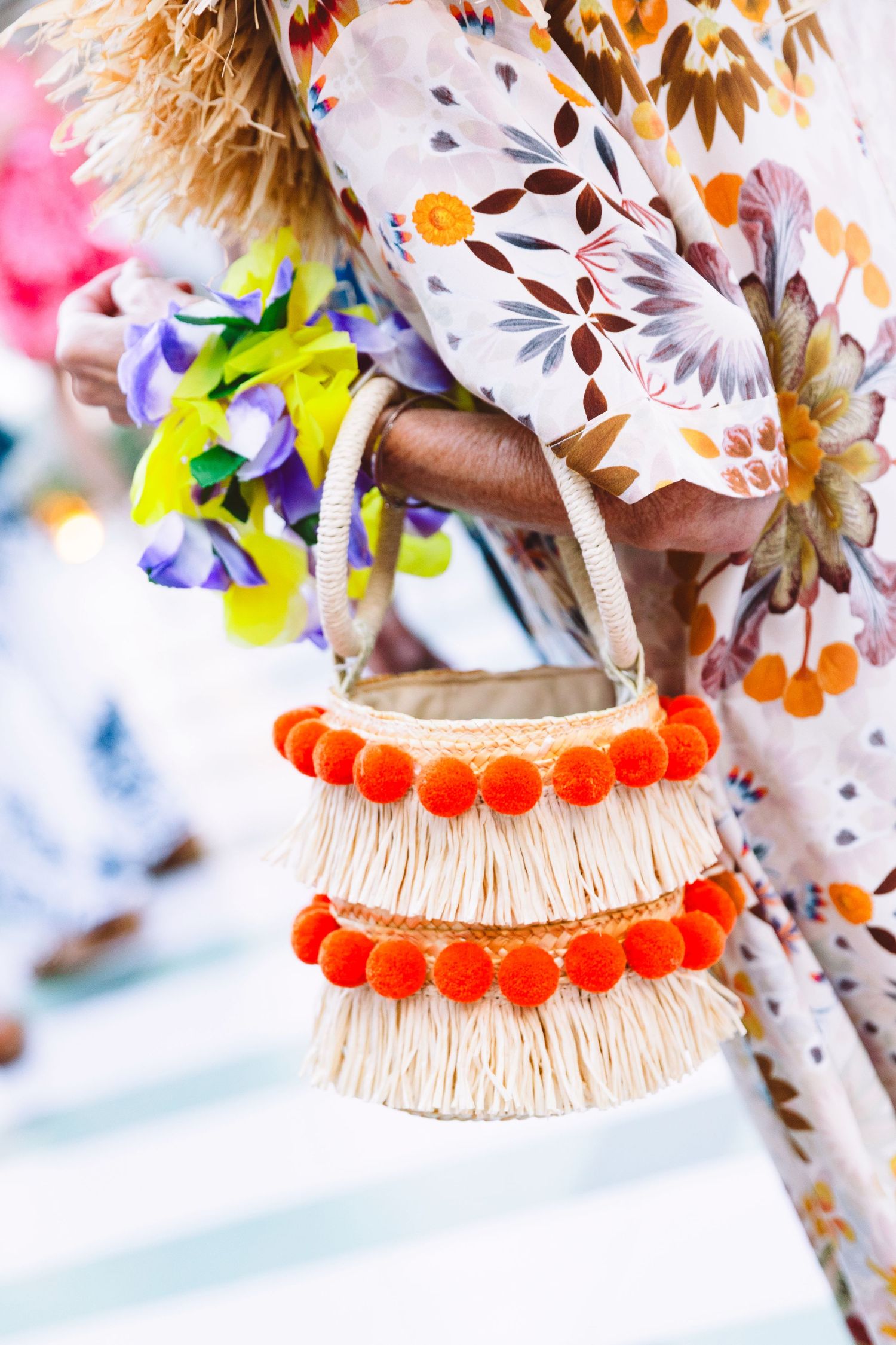 Person carrying a straw bag with orange pom-poms and colorful flowers, wearing a floral print dress.