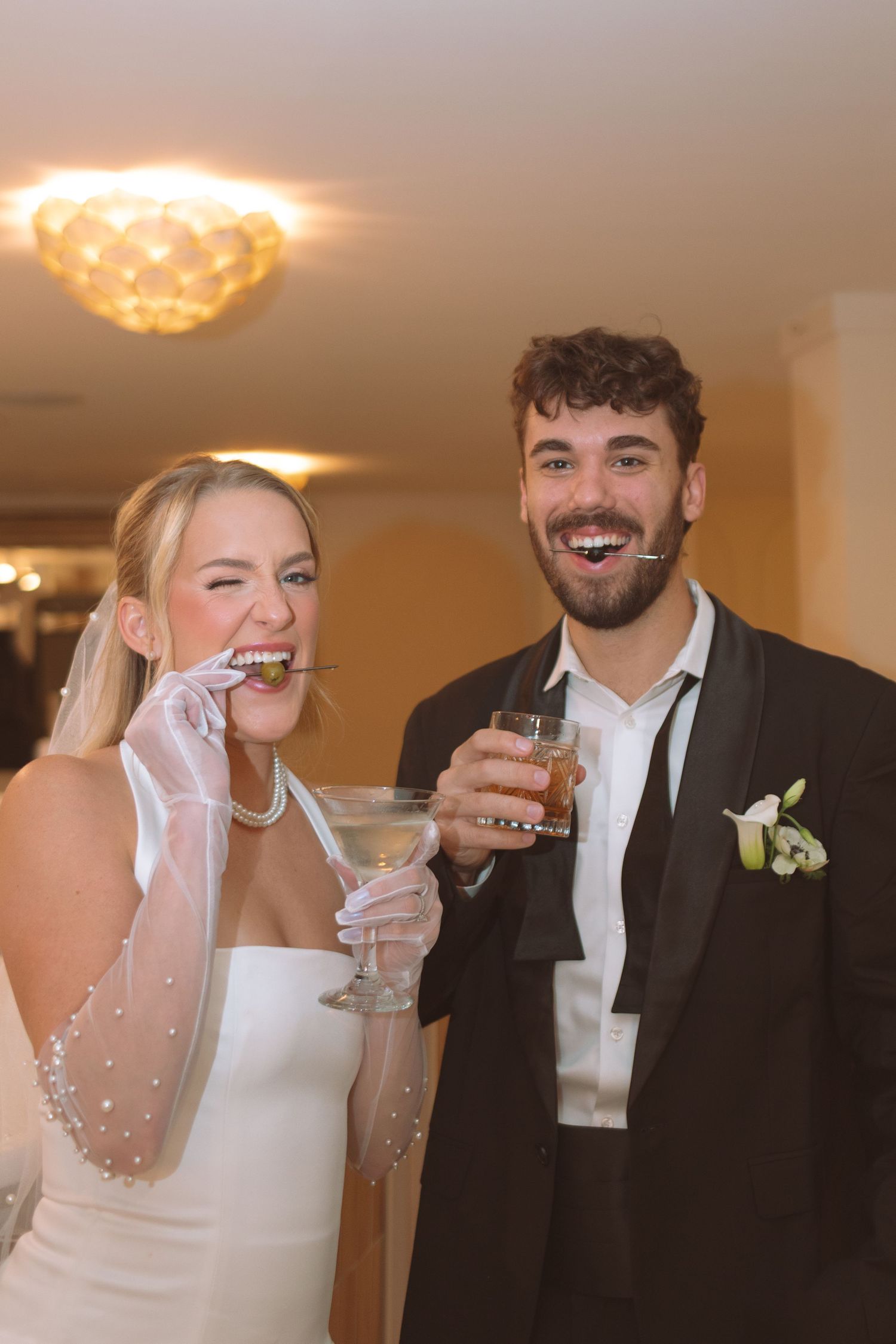 Laughing bride and groom holding cocktails at wedding reception, bride wearing veil and pearl gloves.