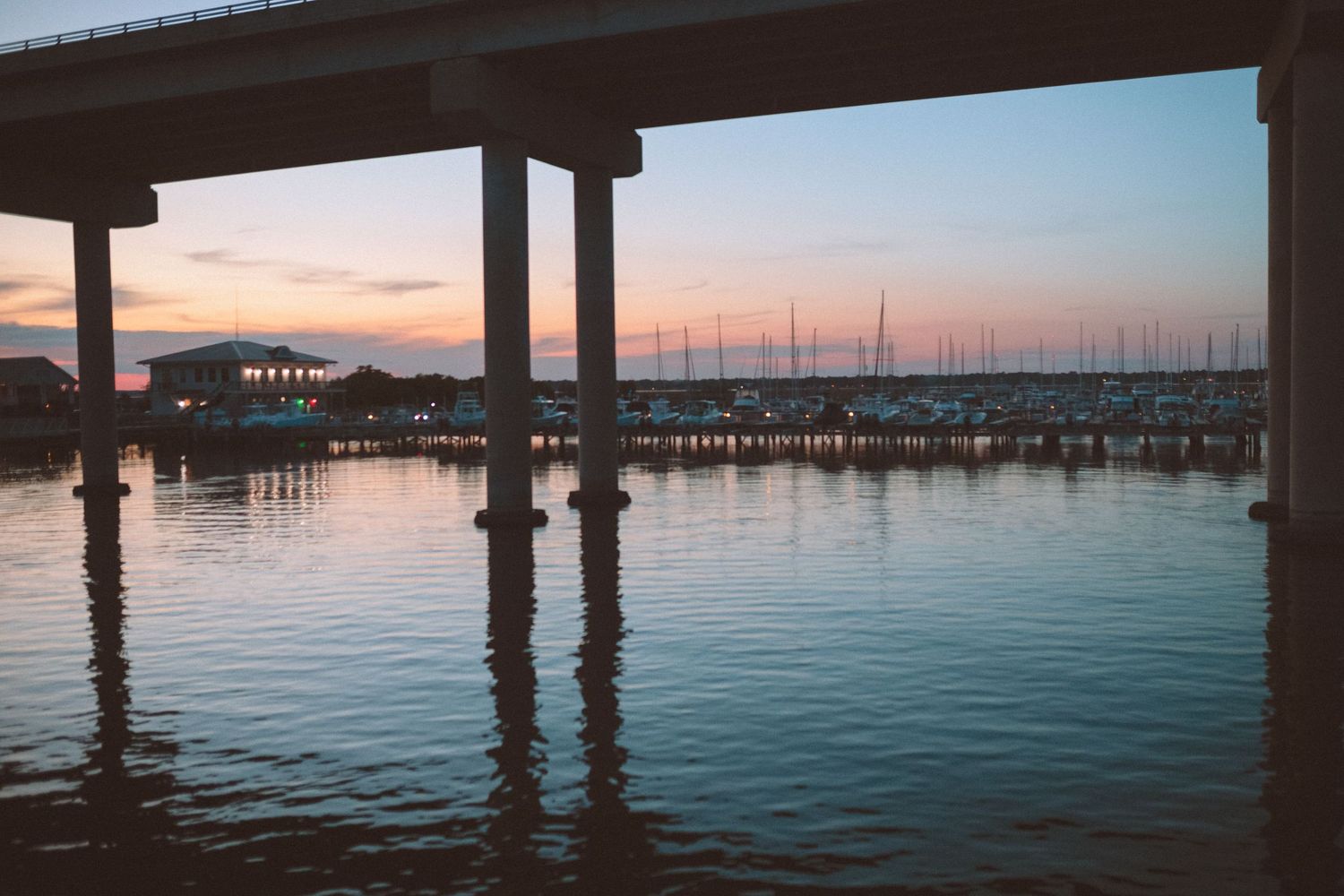 A serene marina at sunset viewed from under a bridge with calm reflective waters and sailboats in the background.