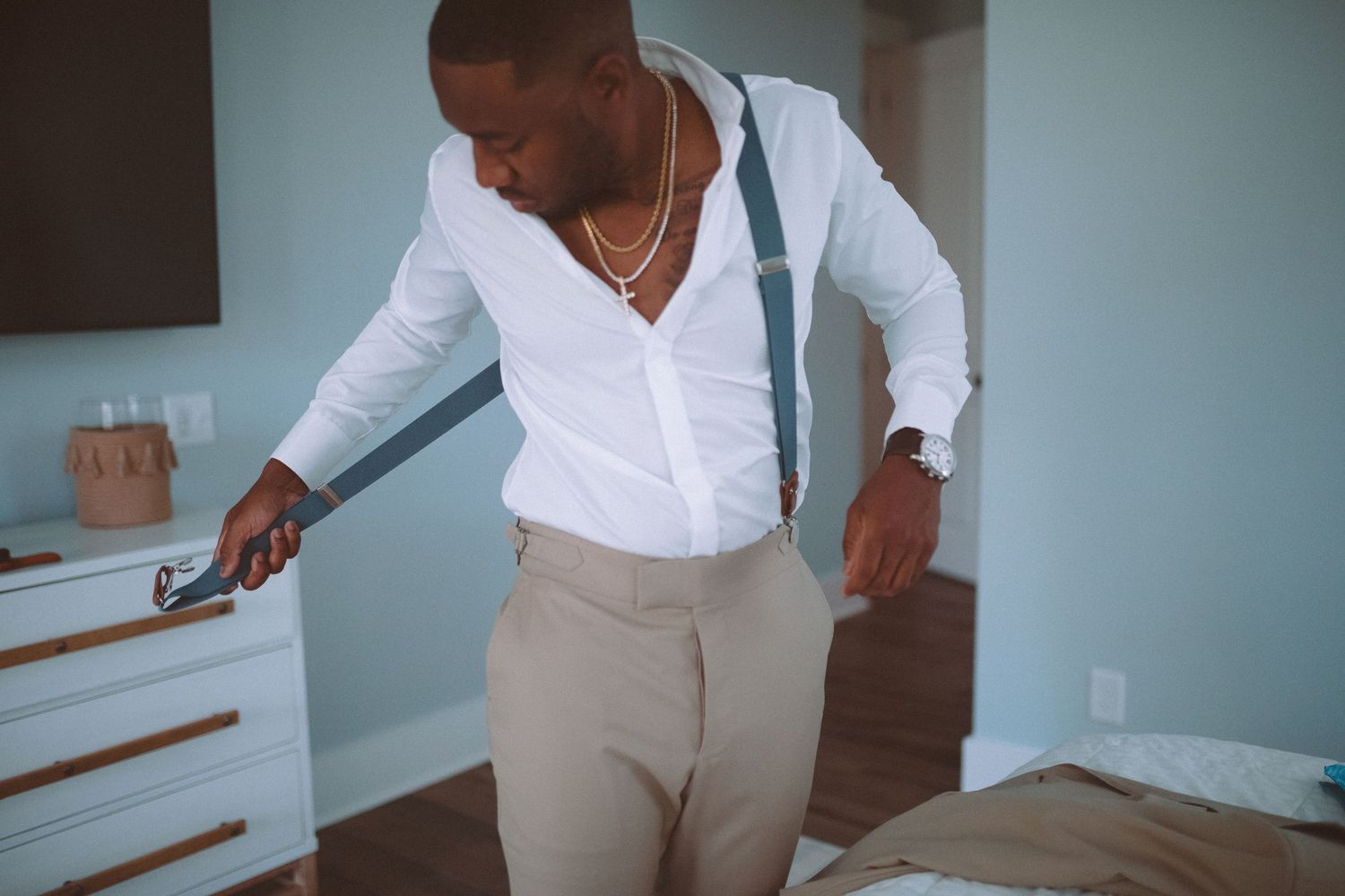 Groom in white shirt and khaki pants adjusting his suspenders while getting ready in a bedroom.