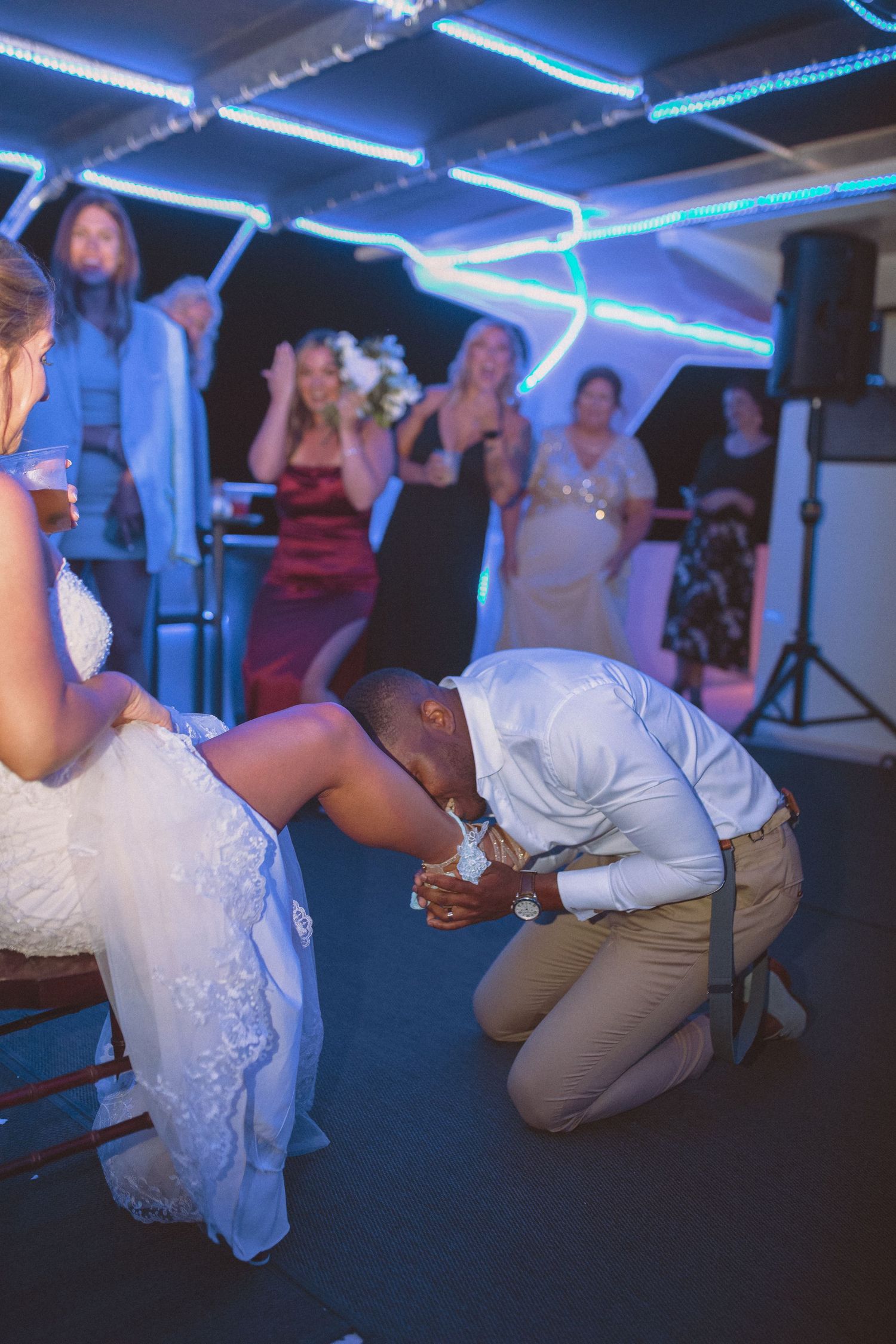 Groom kneeling before seated bride during garter removal at wedding reception celebration.