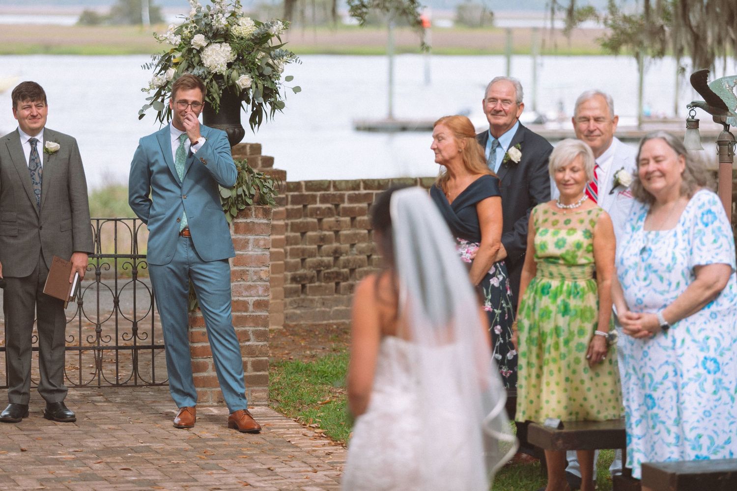 Emotional groom wipes tears watching bride walk down aisle at outdoor waterfront wedding ceremony.