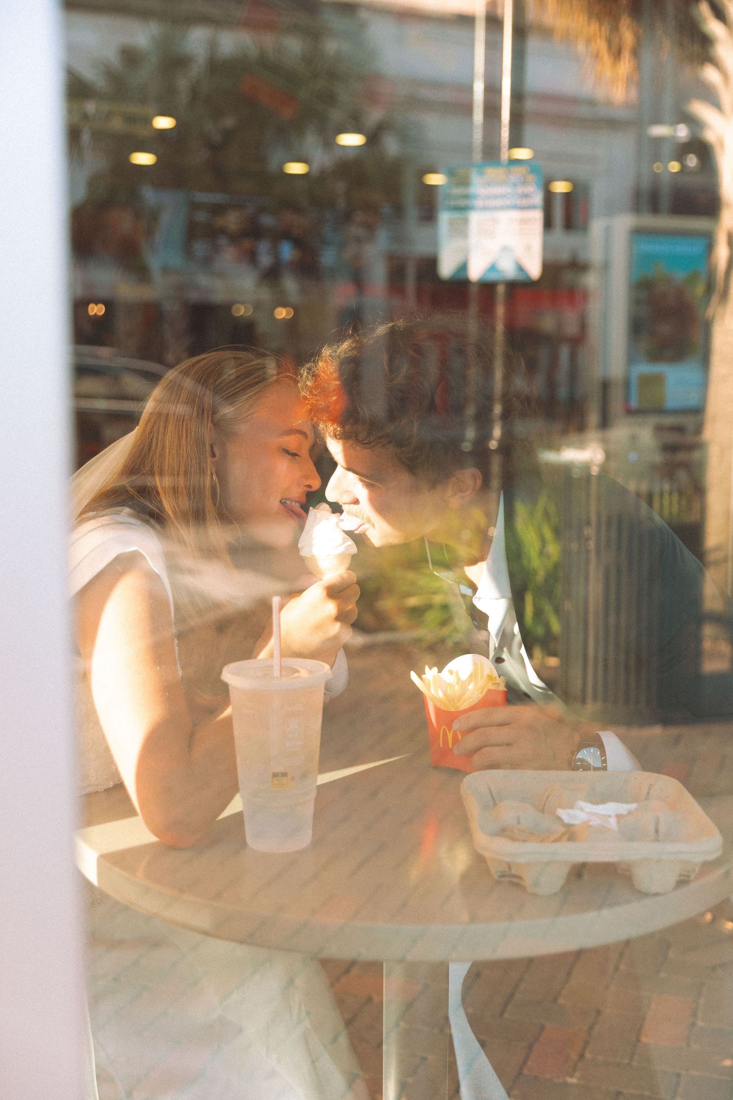 Bride and groom sharing a romantic moment while eating at a restaurant, photographed through a window.