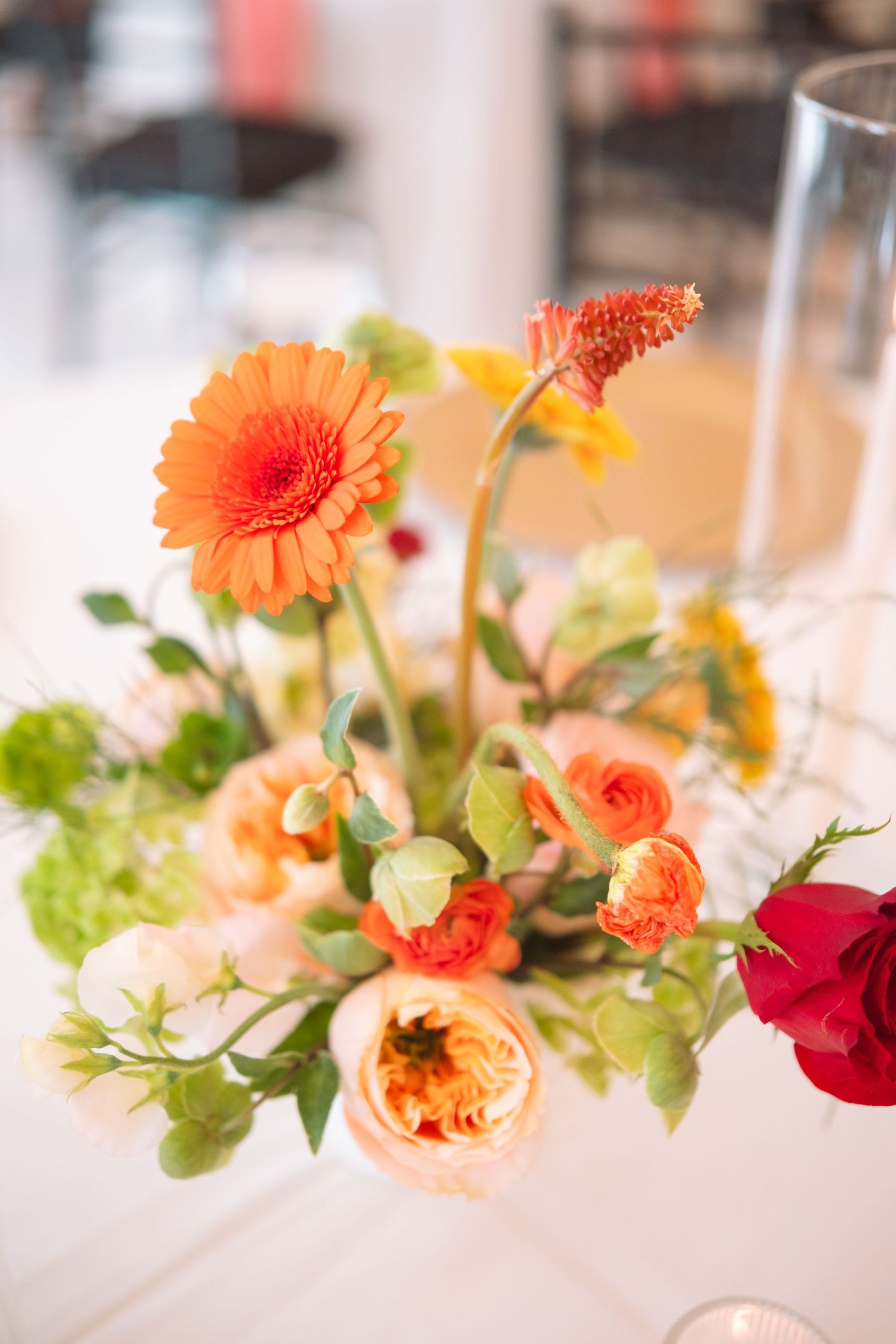 Close-up of orange gerbera daisy and peach roses centerpiece with green foliage on white table.