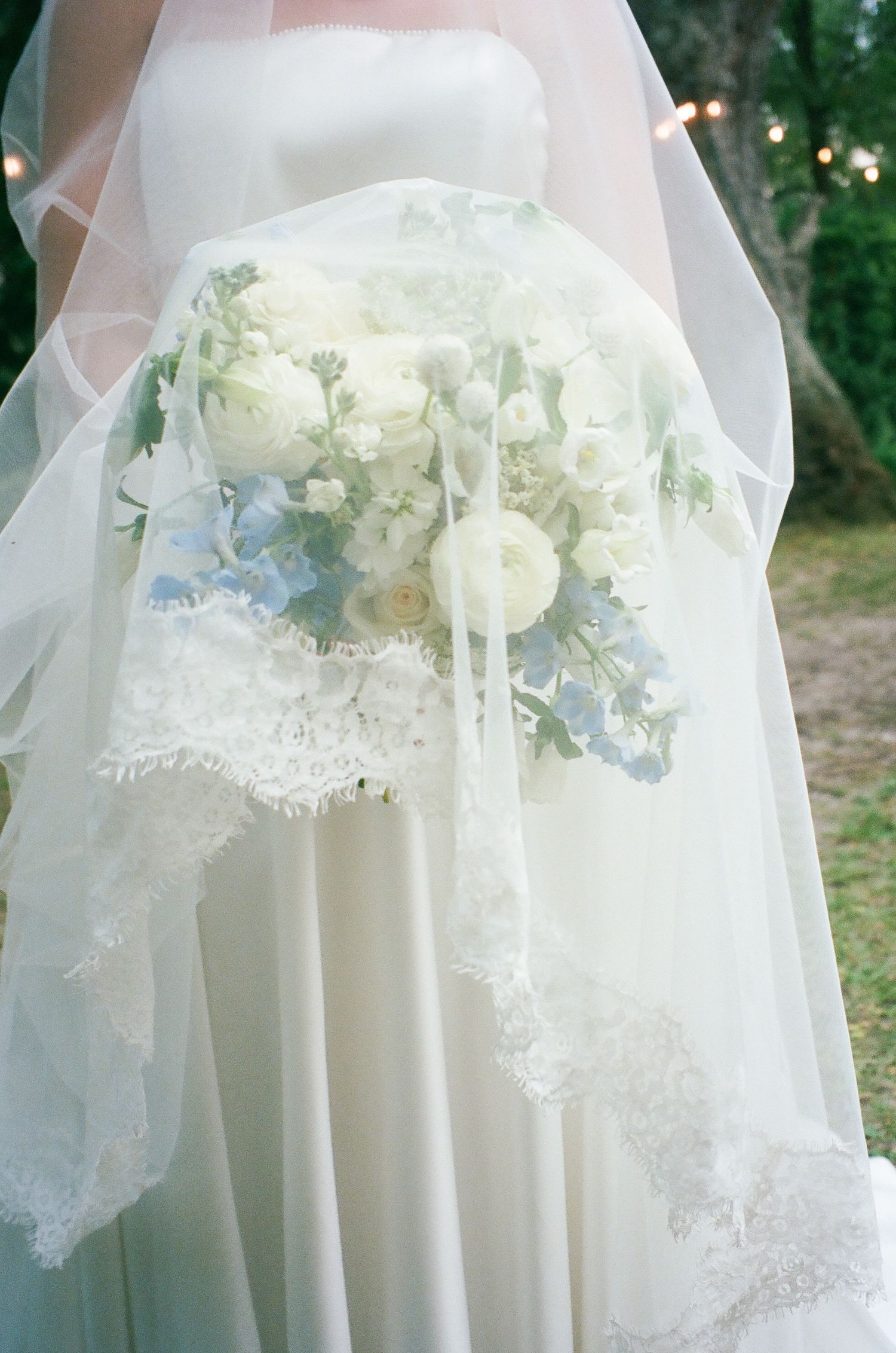 Bride in white strapless gown holding elegant bouquet of white ranunculus and soft blue hydrangeas under a lace-trimmed veil.