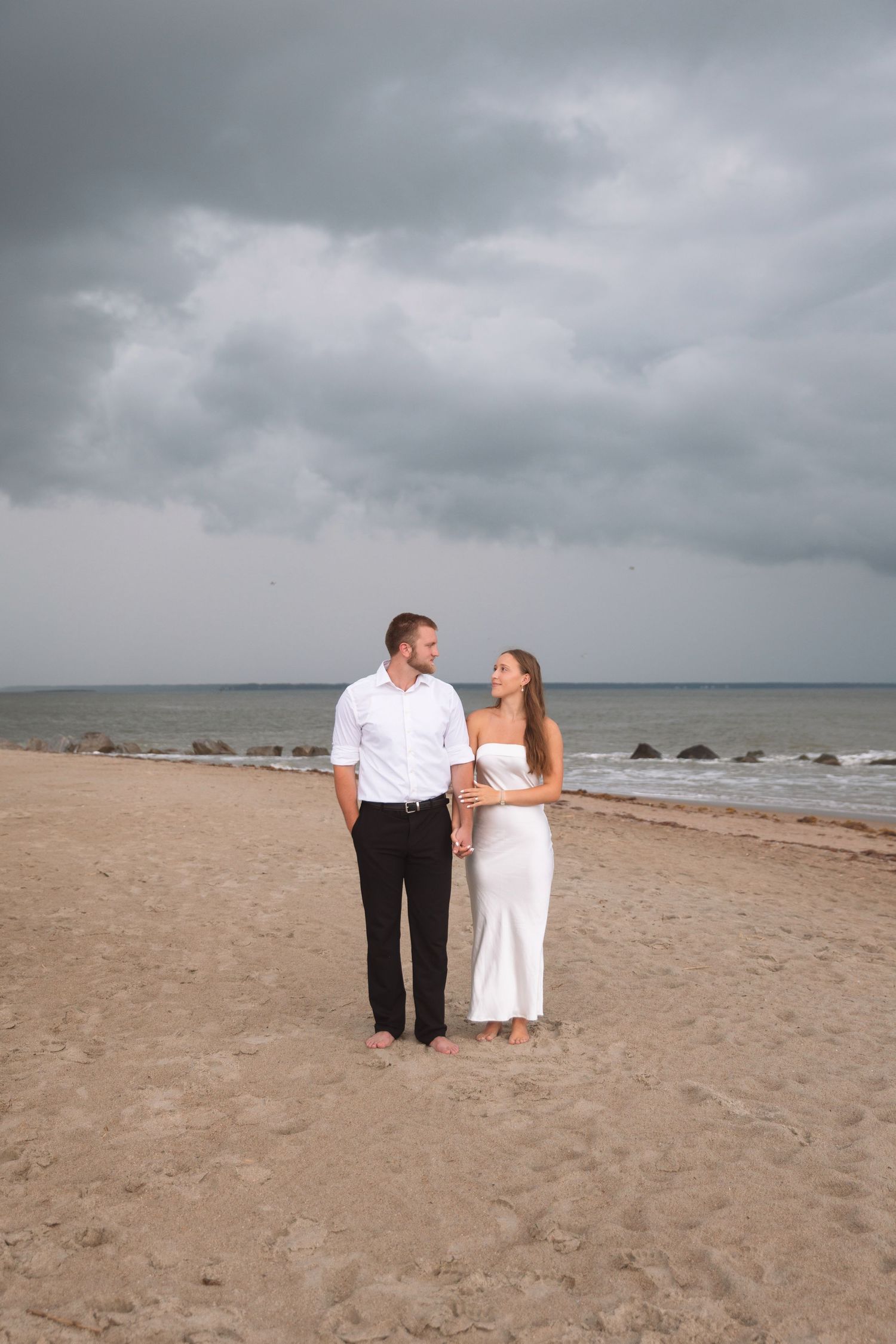 Couple in white outfits standing barefoot on sandy beach under dramatic cloudy sky.